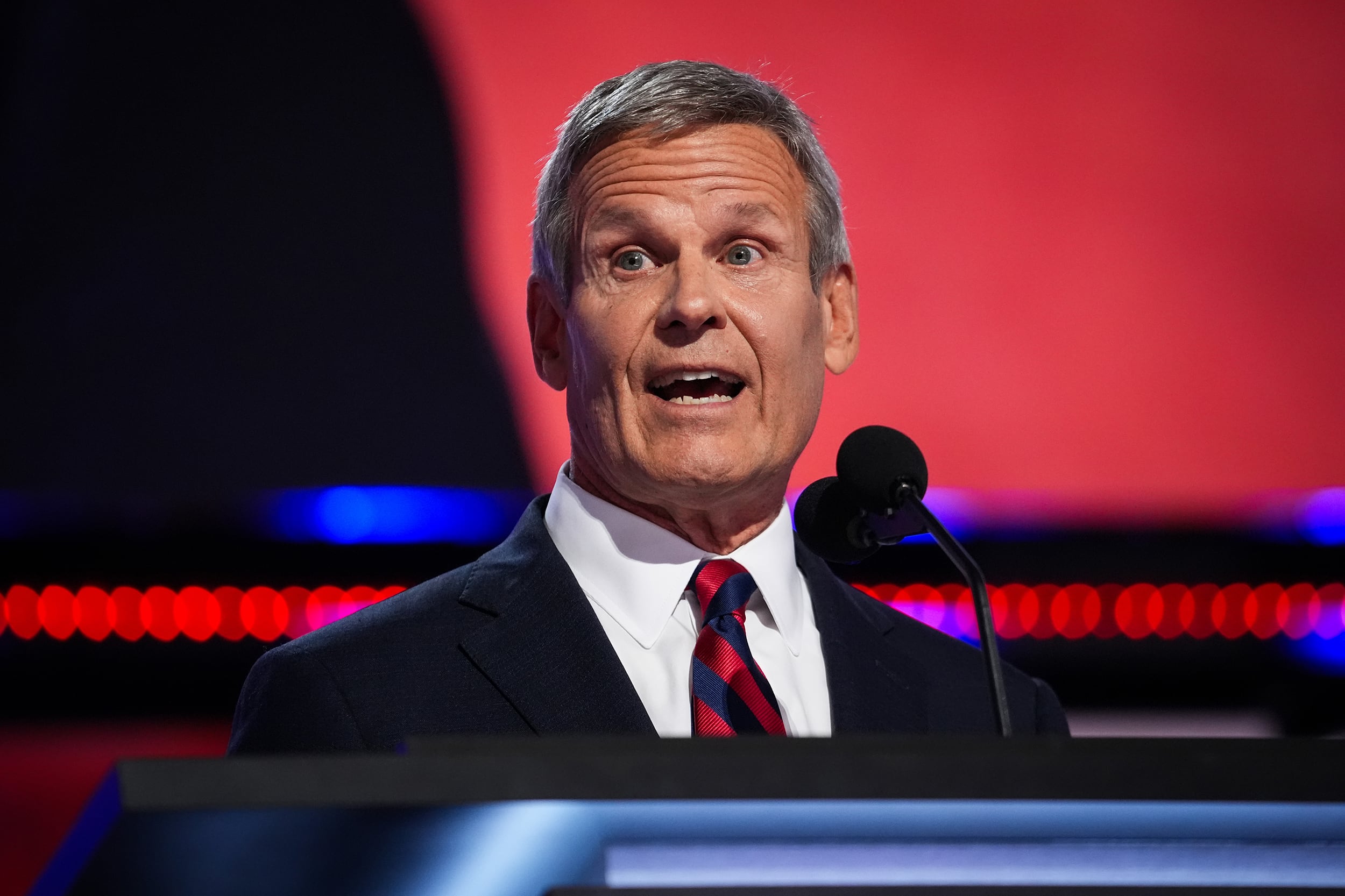 A photograph of a white man in a dark suit with a red tie speaking from behind a podium with a black and red background.