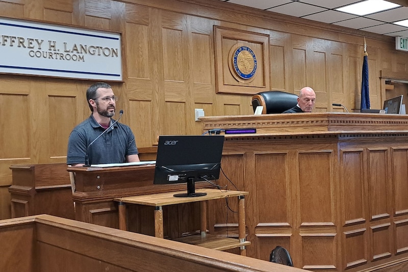 A man in a gray shirt and glasses speaks in a courtroom.