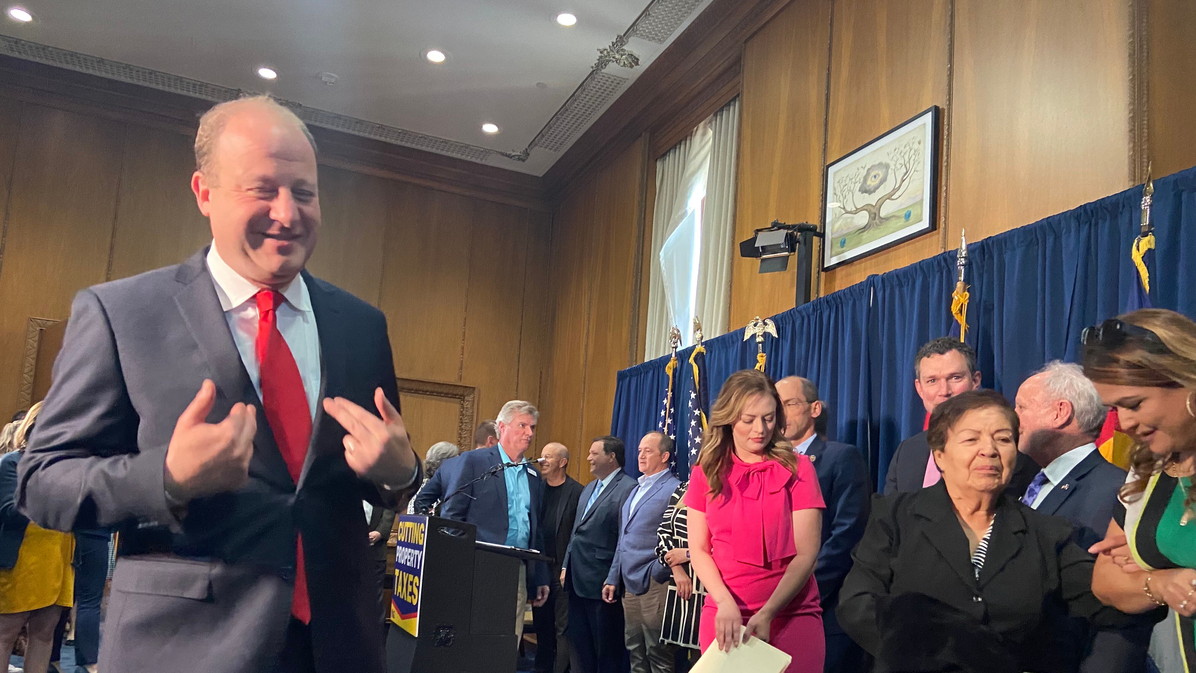 Gov. Jared Polis, standing in a suit and a red tie, seems to point to himself while smiling. Men and women stand, some holding papers, behind him in front of a blue curtain.