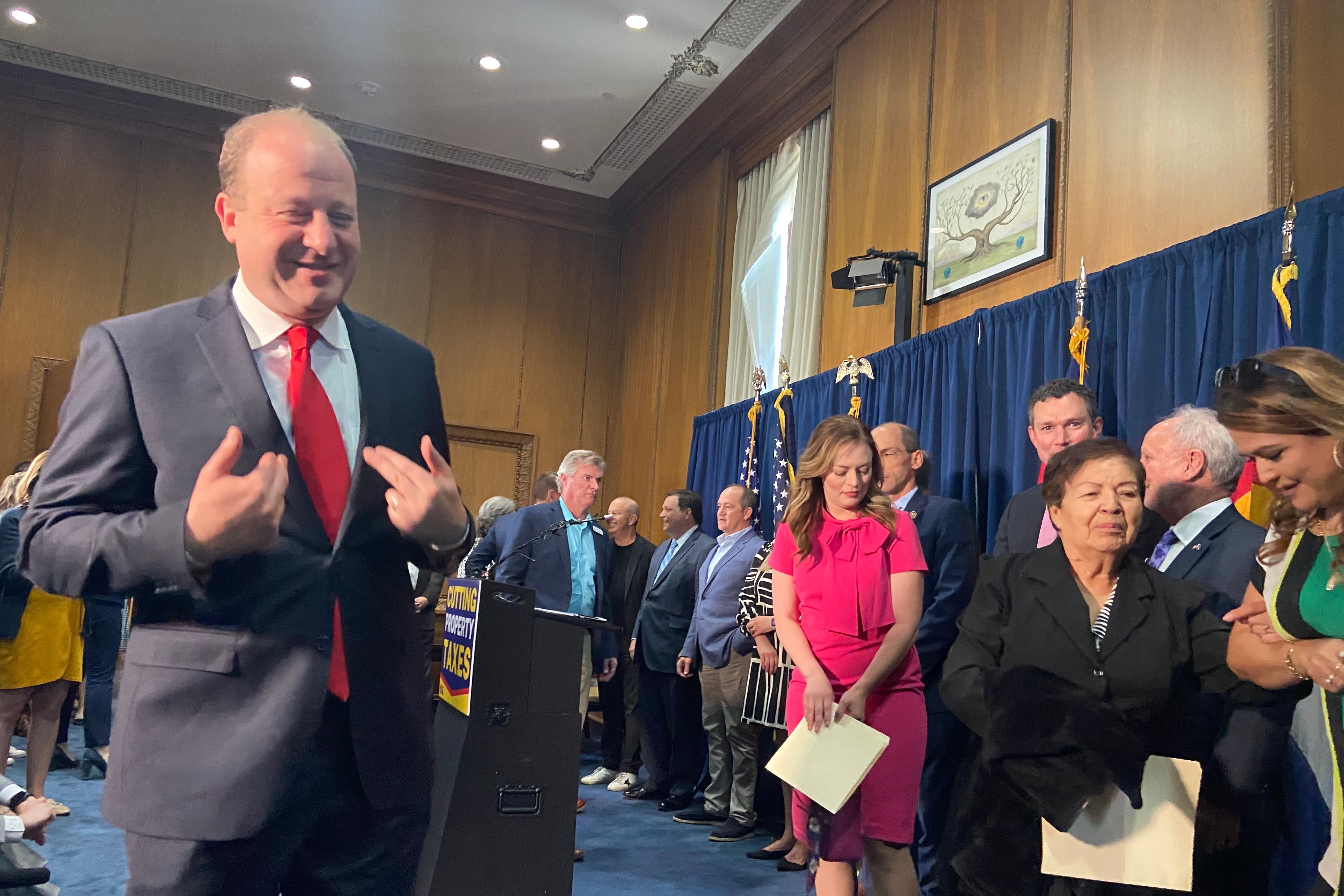 Gov. Jared Polis, standing in a suit and a red tie, seems to point to himself while smiling. Men and women stand, some holding papers, behind him in front of a blue curtain.