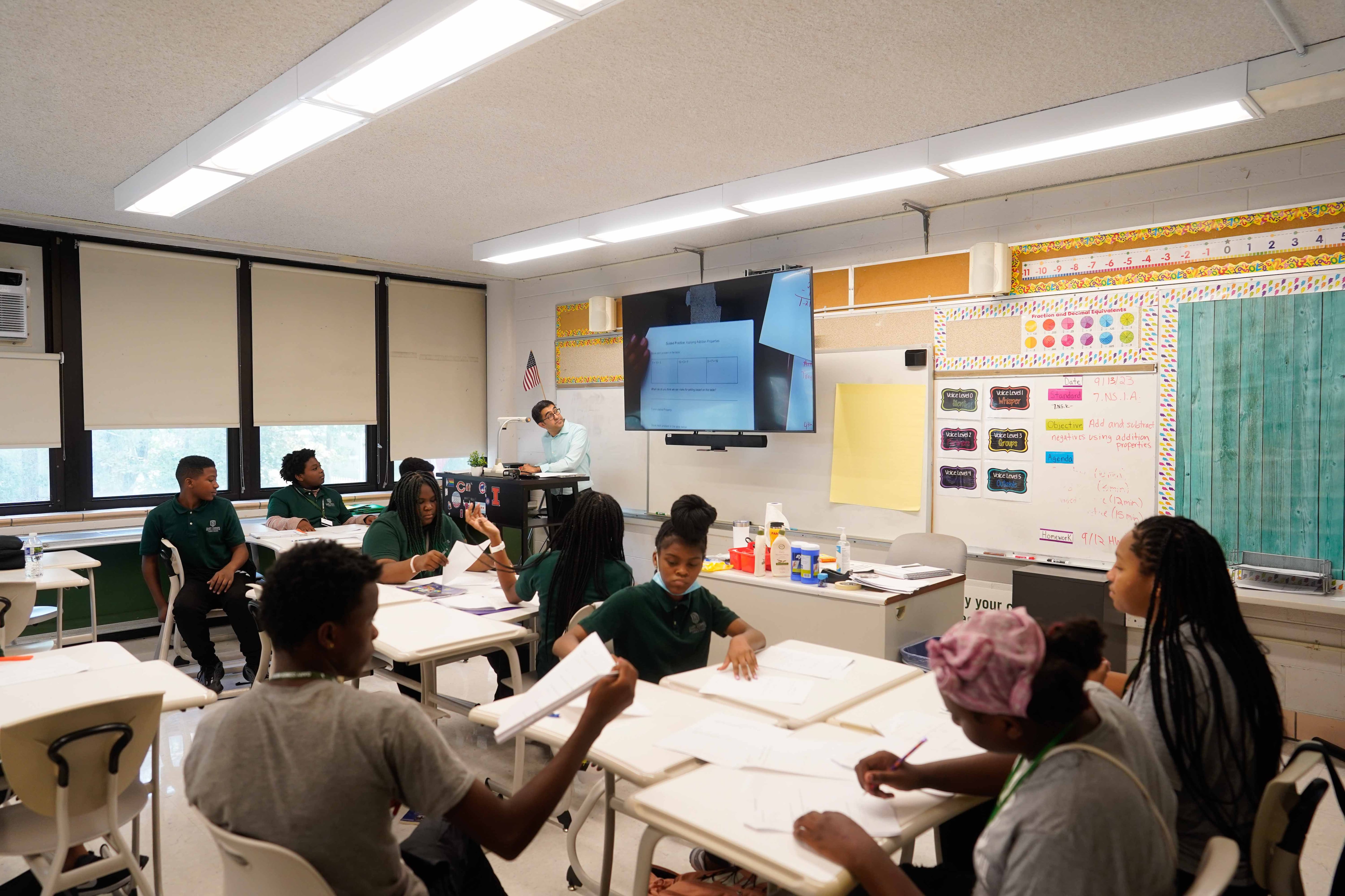 A classroom full of middle school students sitting at their desk while a teacher stands near the dry erase board in a classroom.