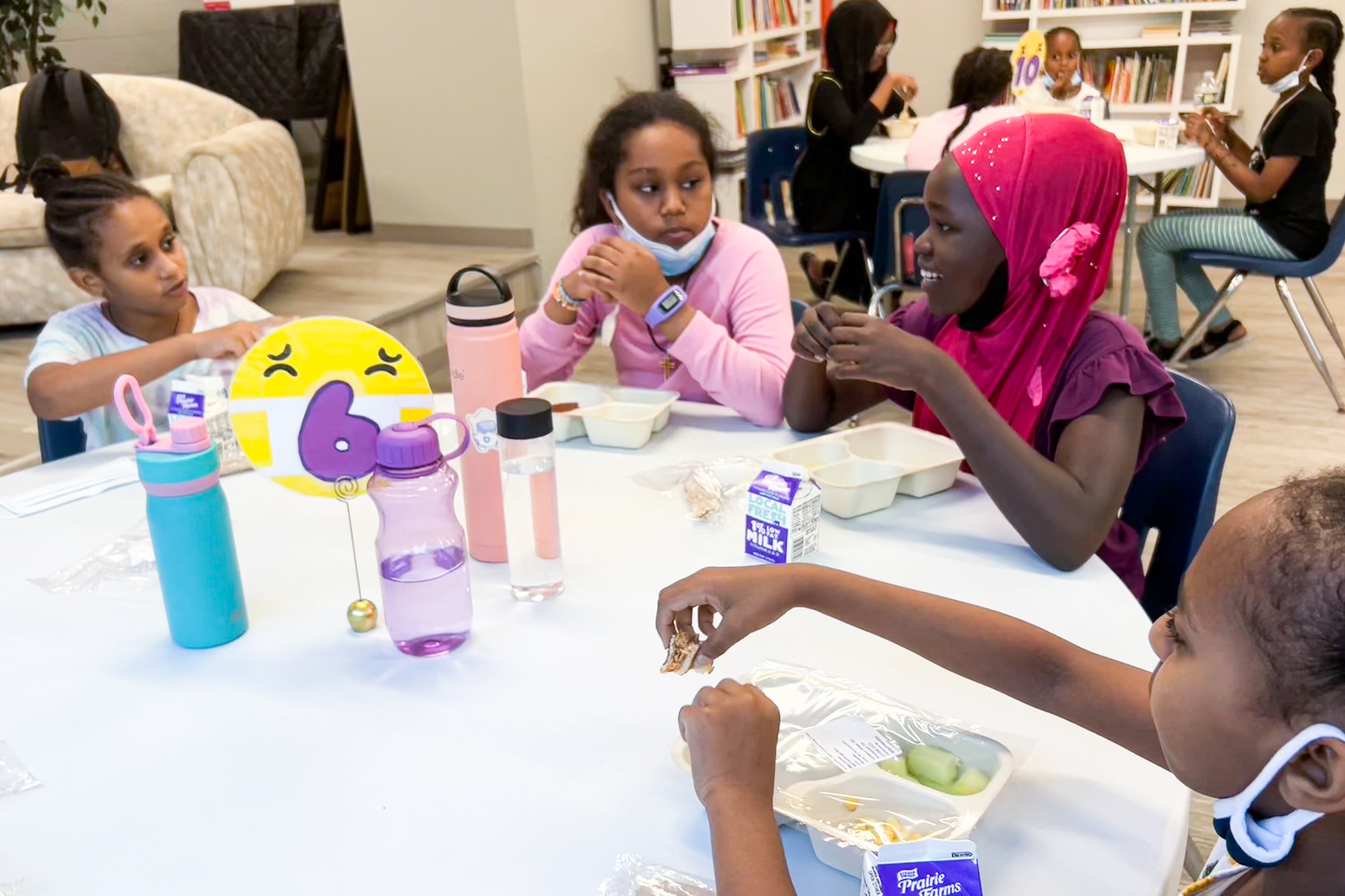 Four children are sitting at a table talking to one another as they eat lunch. Their lunches, milk cartons, and water bottles are all on the table.
