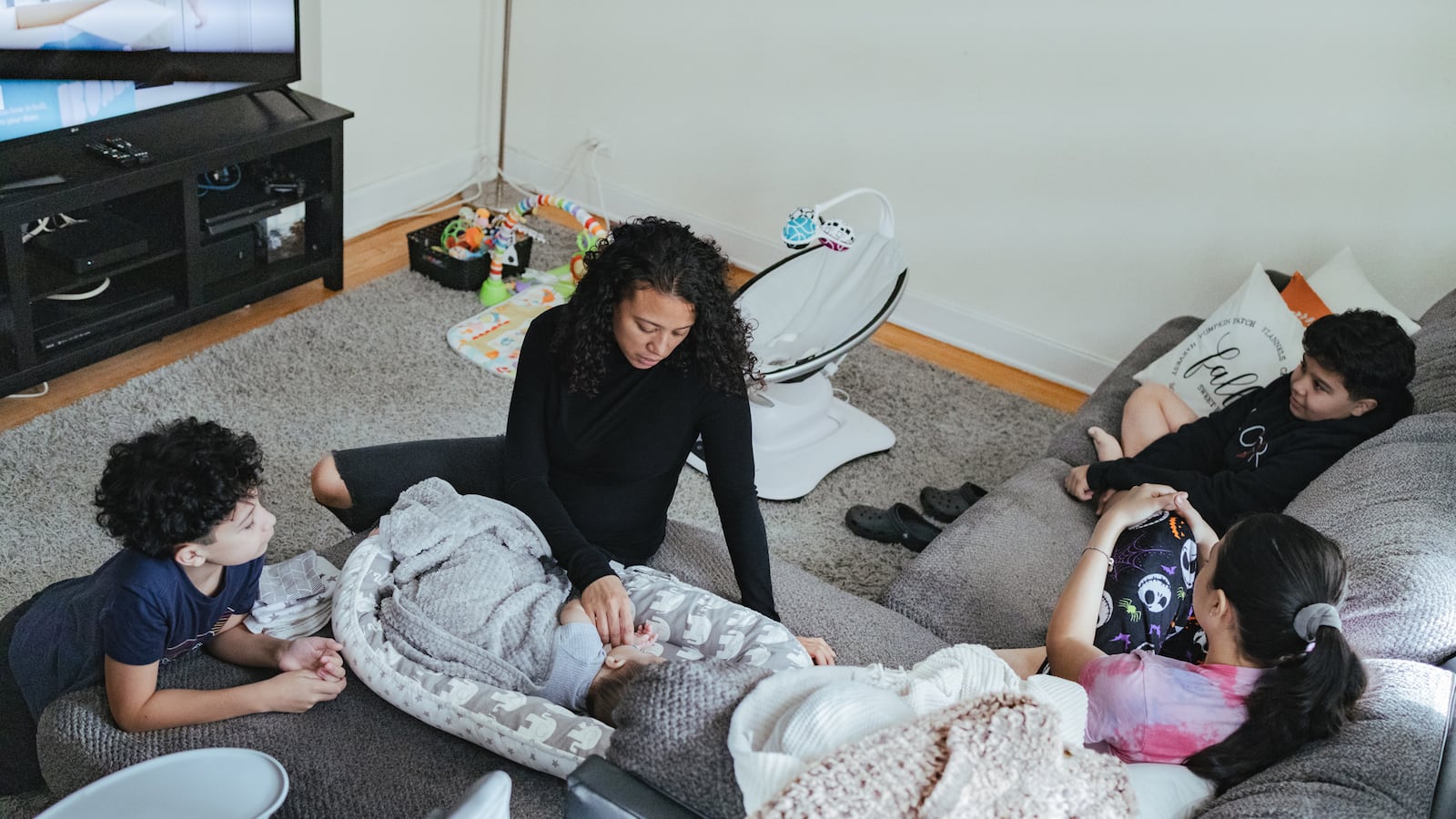 A woman sits with her four children: an infant, two boys and a young girl, in the family’s living room.