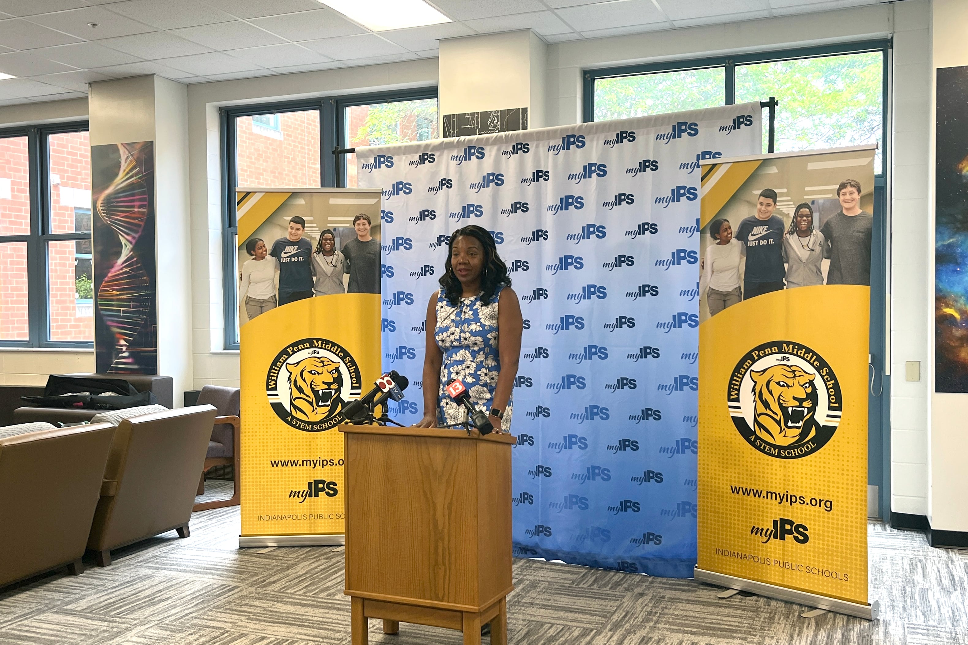 A photograph of a Black woman in a dress standing behind a wooden podium and in front of a background in a school library.