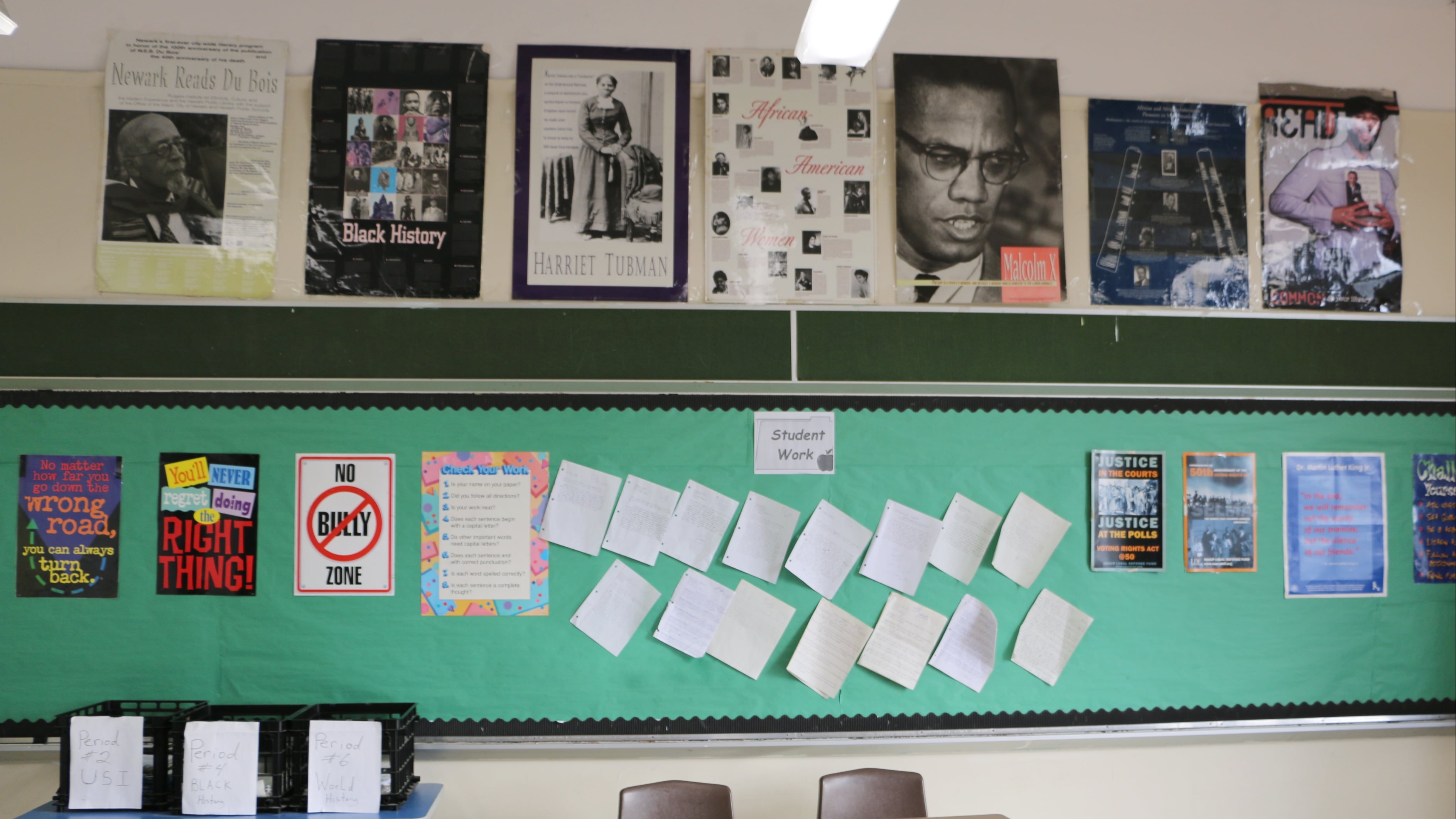 A classroom displays posters of Harriet Tubman, Malcolm X, and others on the wall, along with other instructional materials. Several desks and a small table are also visible.
