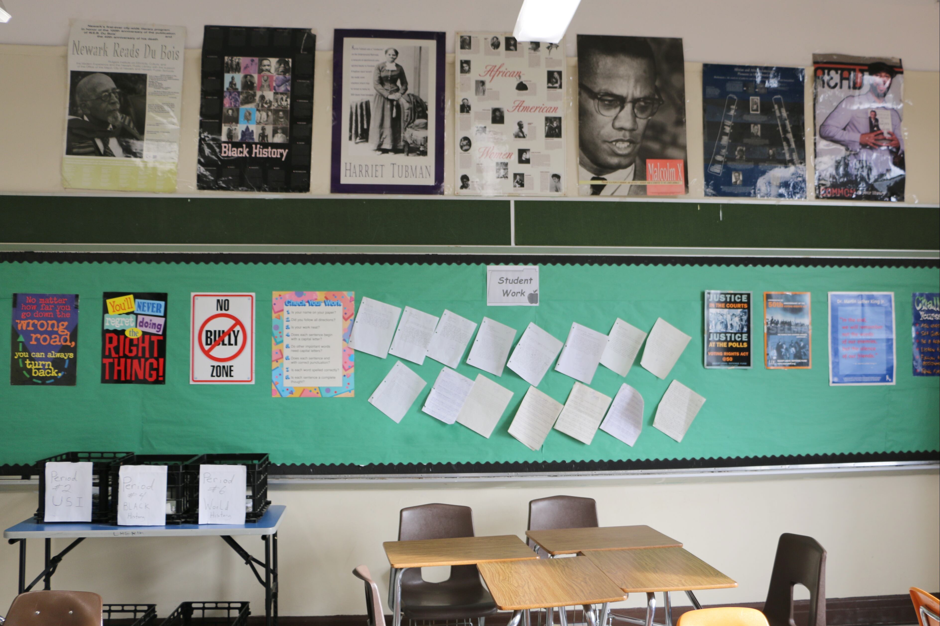 A classroom displays posters of Harriet Tubman, Malcolm X, and others on the wall, along with other instructional materials. Several desks and a small table are also visible.