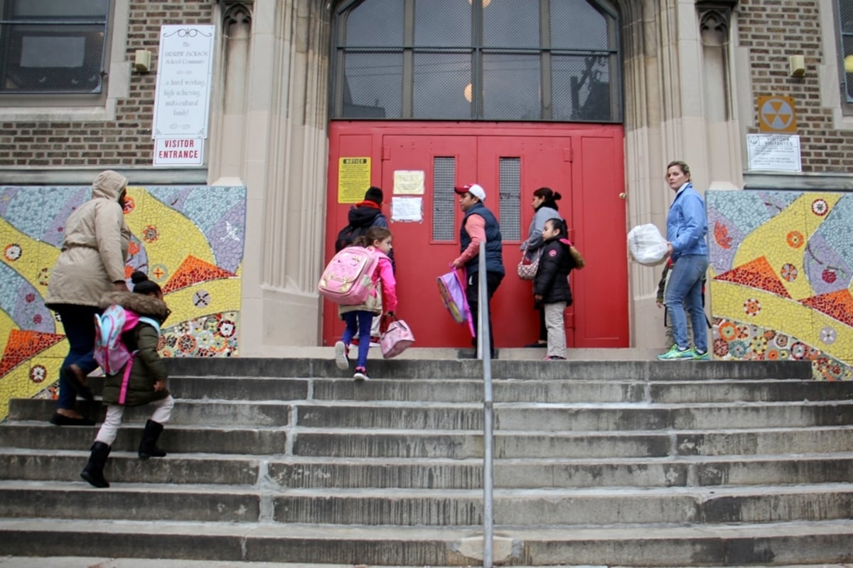 Children and adults stand on stairs in front of a set of red double doors with glass windows above it.