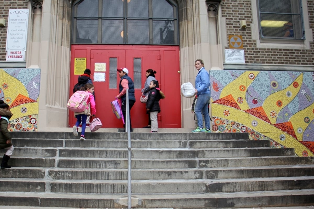Children and adults stand on stairs in front of a set of red double doors with glass windows above it.