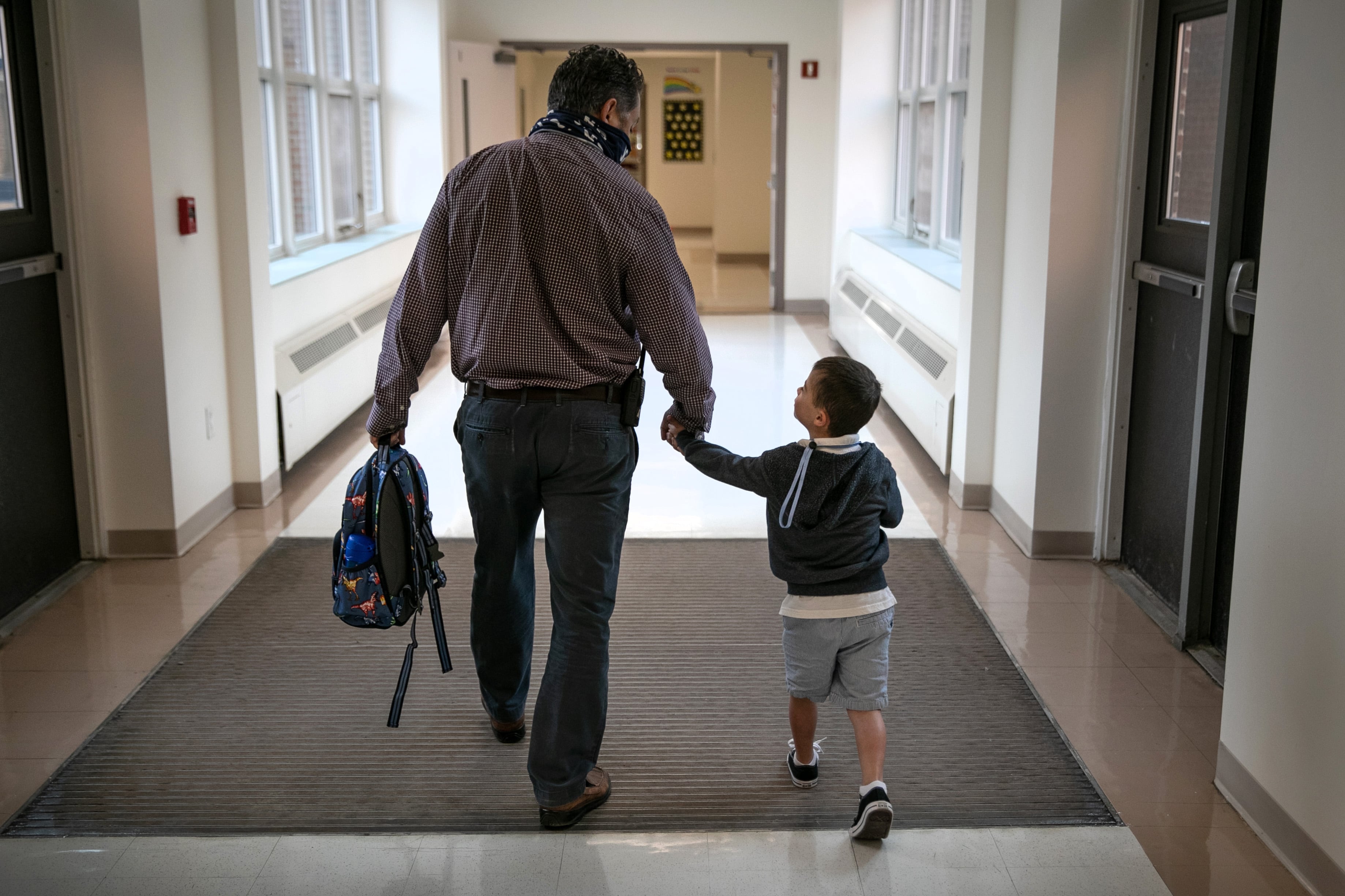 A man walks while holding hands with a young boy in a school hallway, grasping the child’s backpack in the other hand.