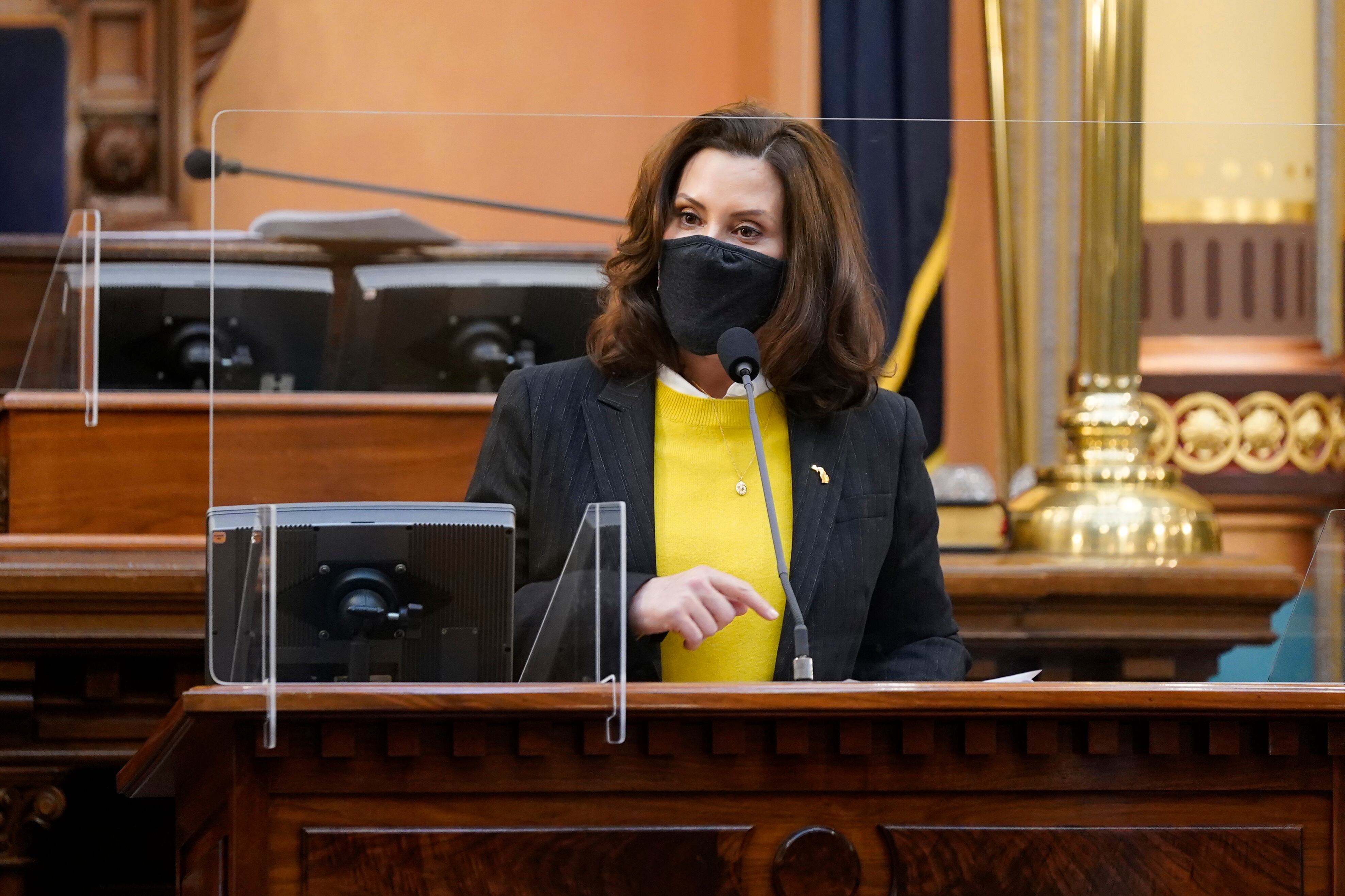 Michigan Governor Gretchen Whitmer, wearing a black coat, yellow sweater and protective mask, speaks behind a partition in the Michigan State Capitol.