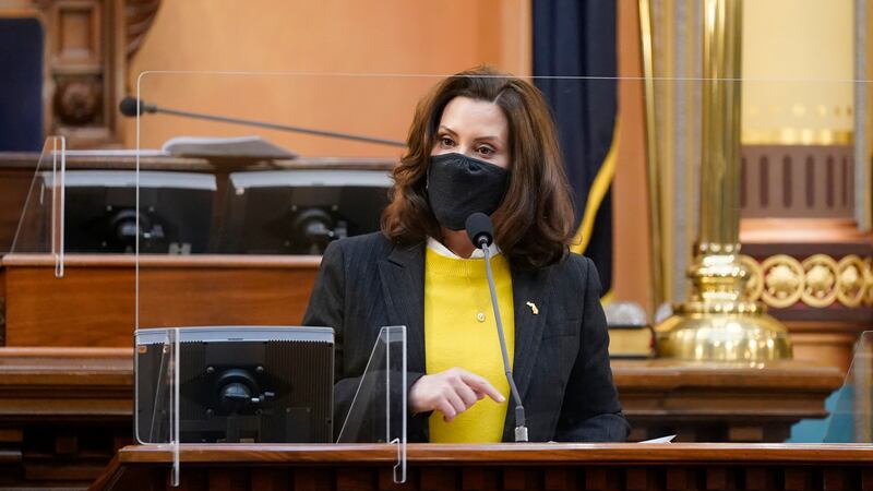 Michigan Governor Gretchen Whitmer, wearing a black coat, yellow sweater and protective mask, speaks behind a partition in the Michigan State Capitol.