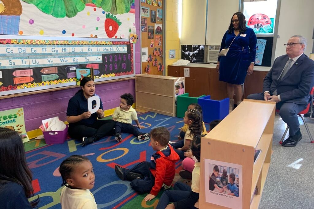 A person sits on the floor and holds up the letter “D” as a child sits on the floor and other children and adults look on in a classroom.