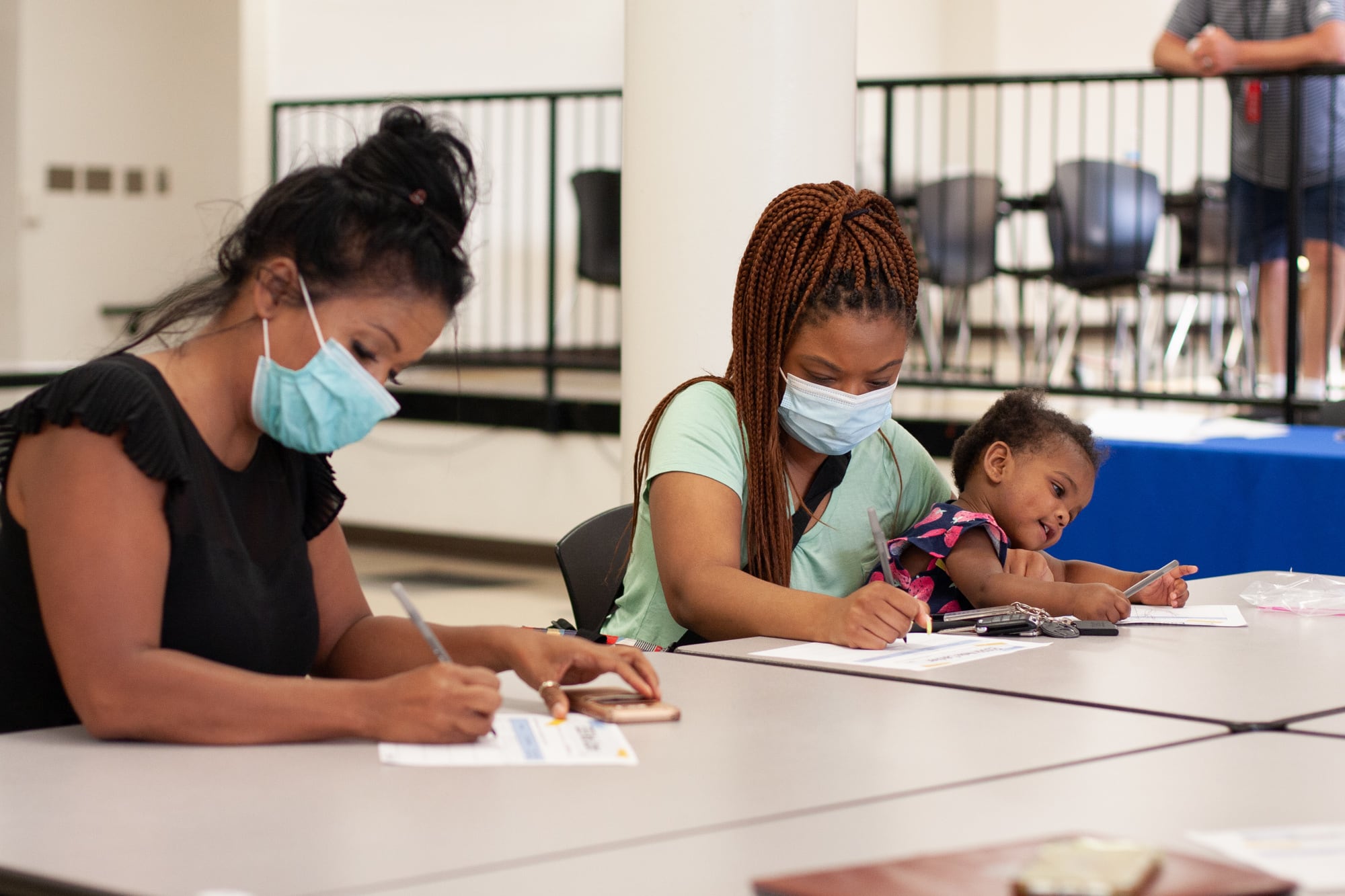 A woman in a black shirt wearing a blue face mask with her hair in a bun and a woman in a teal shirt with a blue face mask holding a distracted young child on her lap sit at a table and write on pieces of paper.