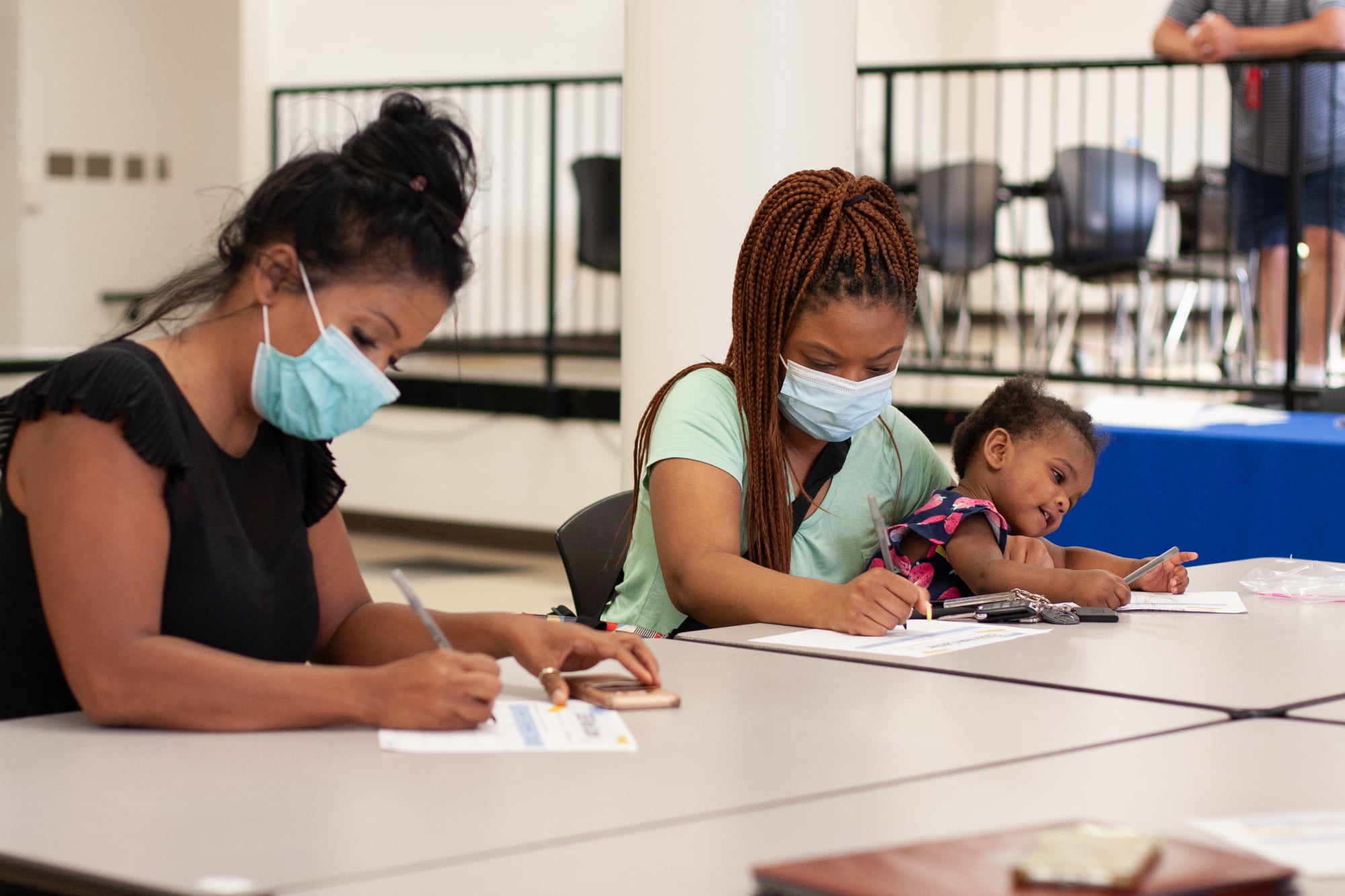 A woman in a black shirt wearing a blue face mask with her hair in a bun and a woman in a teal shirt with a blue face mask holding a distracted young child on her lap sit at a table and write on pieces of paper.