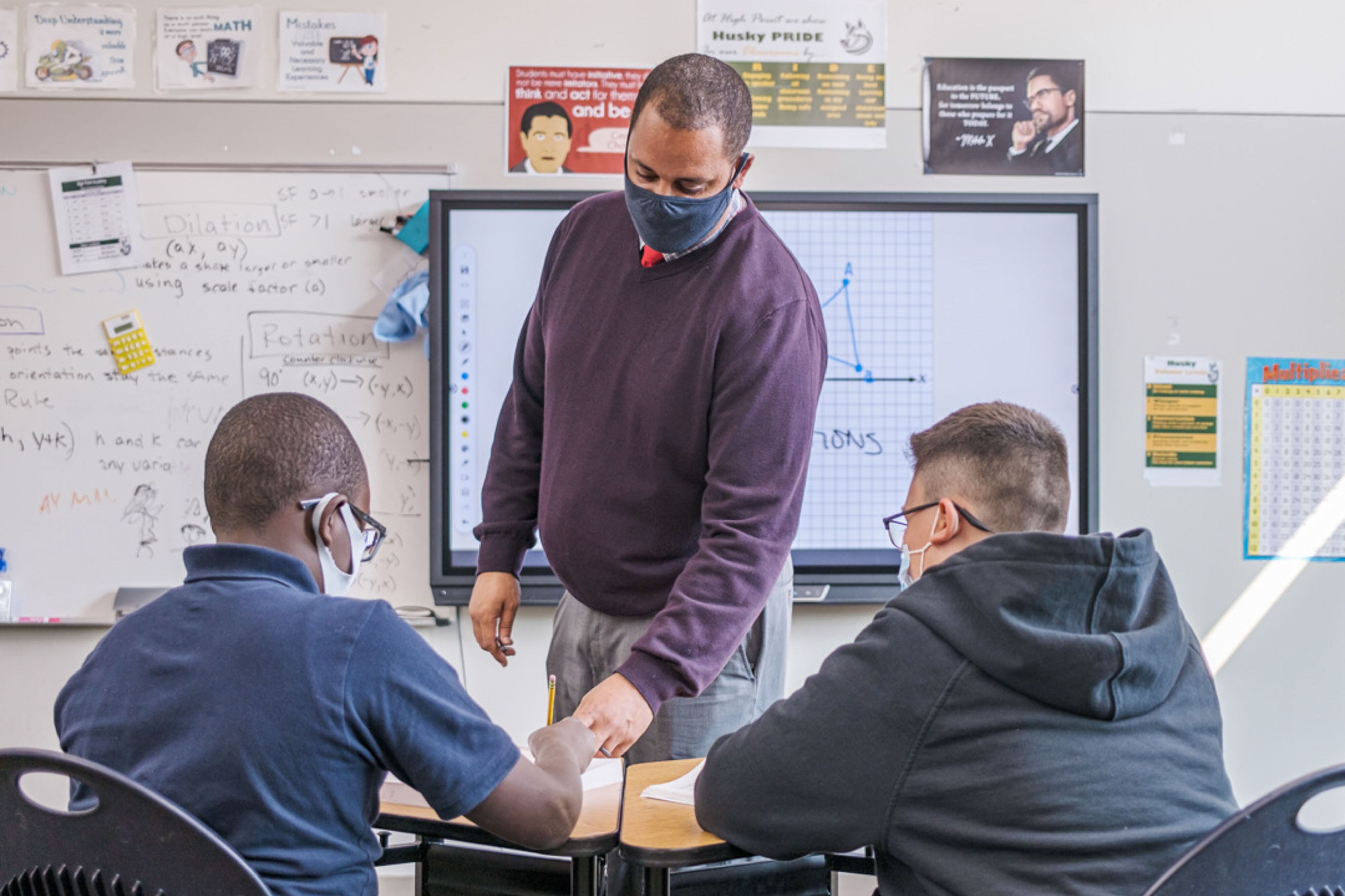 Denver School Board candidate Gene Fashaw, standing in a classroom and wearing a mask, works with two students sitting at their desks in a classroom.
