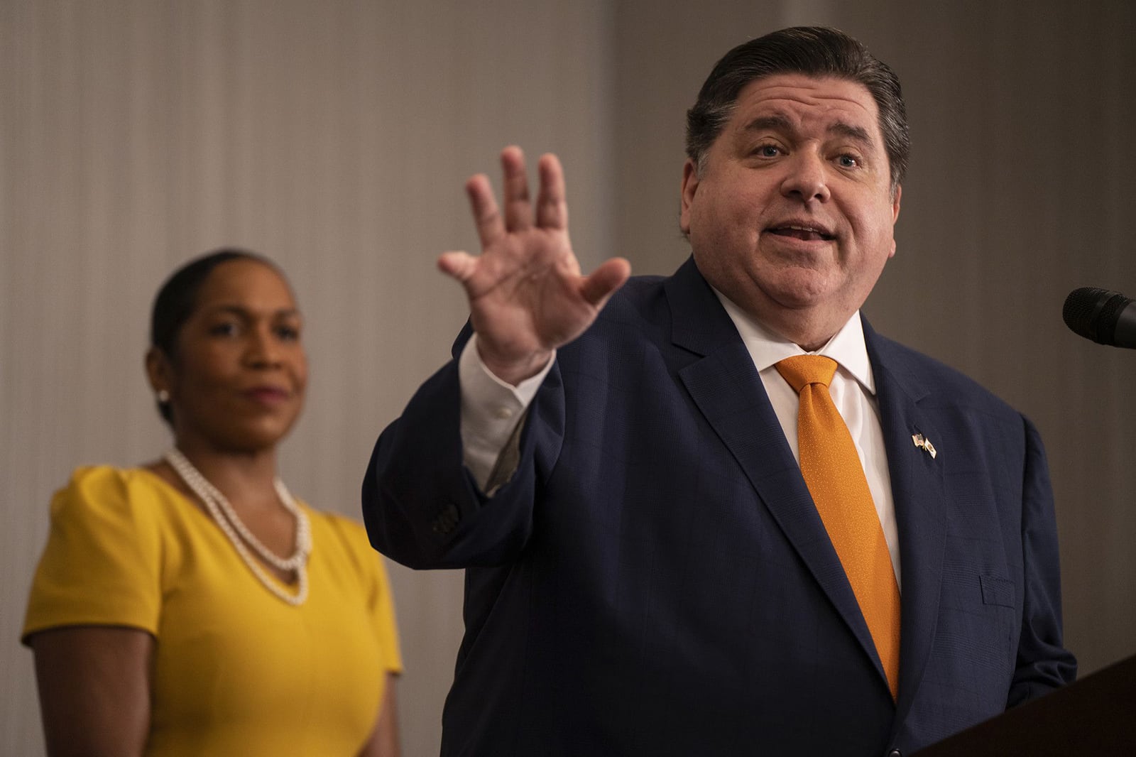 Illinois Gov. J.B. Pritzker is speaking at a microphone, facing the camera with his hand in the air. Lt. Gov. Juliana Stratton is seen in the background look at Pritzker.