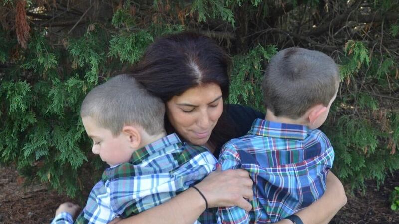 A mother with brown hair holds her two twin sons, both have buzzed hair and are wearing flannel shirts.