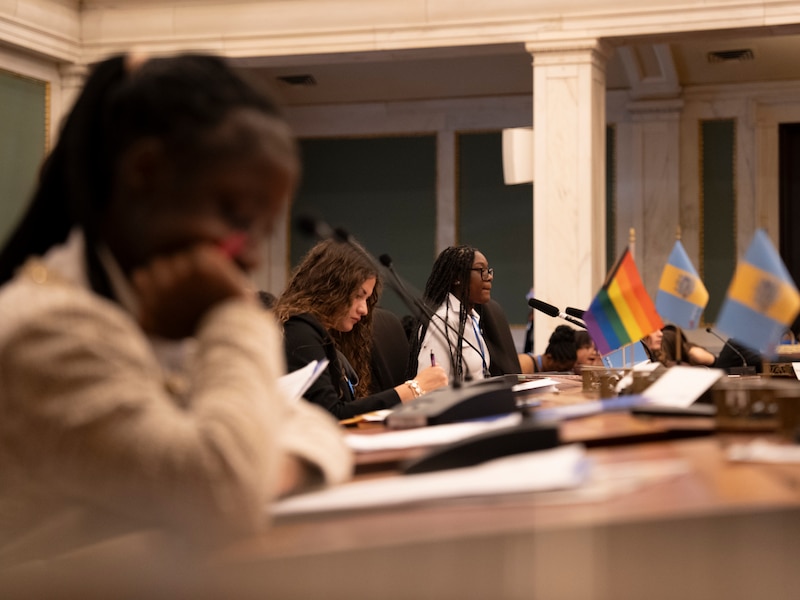 A photograph of three teenage women sitting at desks; one is reading, another is writing.