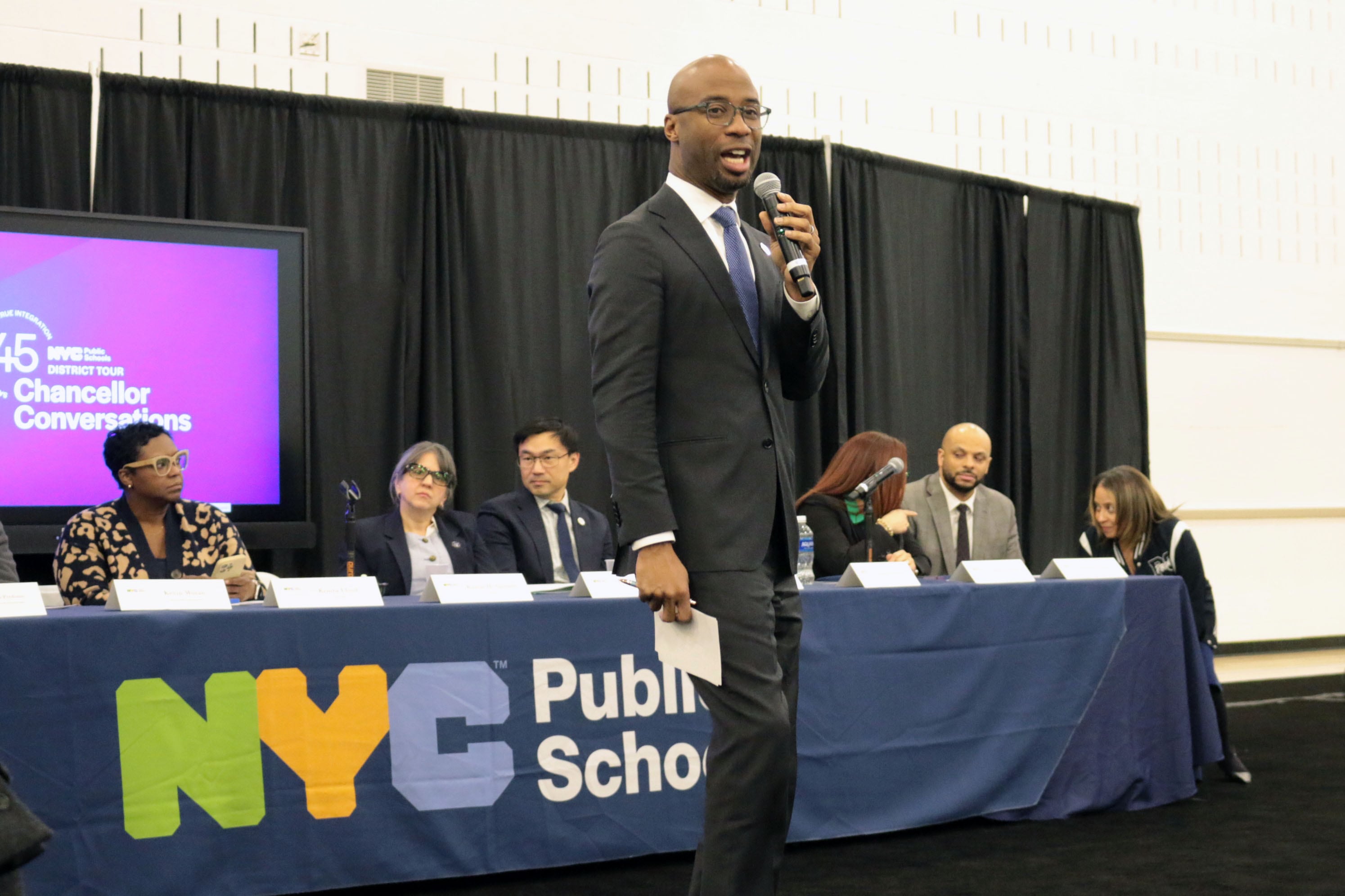 A photograph of a Black man in suit and tie holding a microphone speaking in front of crowd with a group of people sitting in a row of chairs and a table in the background.