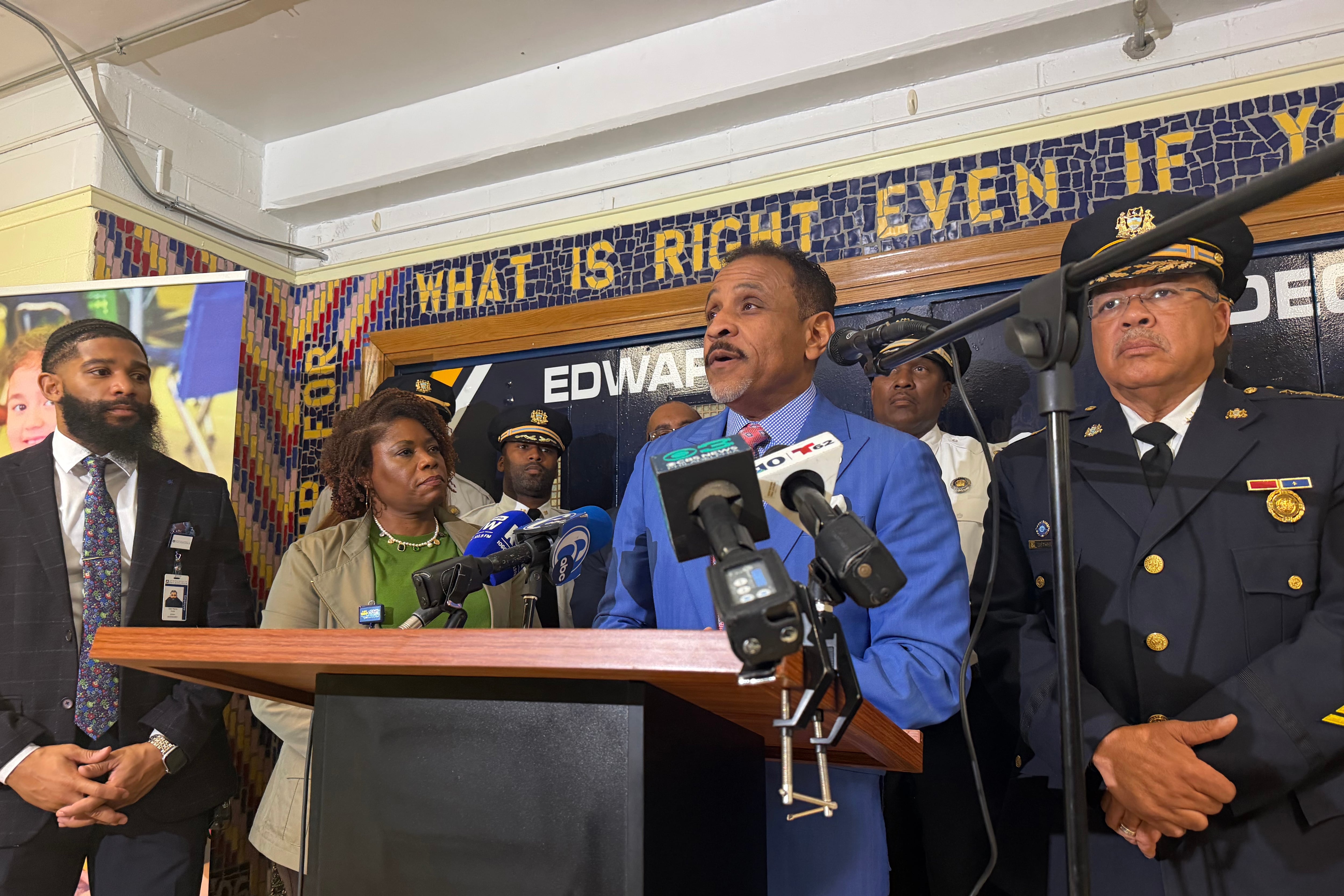 A Black man in a blue suit speaks at a podium with a group of people standing behind him.