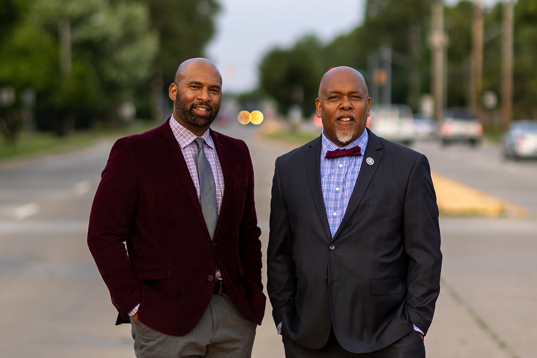 A photograph of two Black men in suits pose together in the middle of a street.