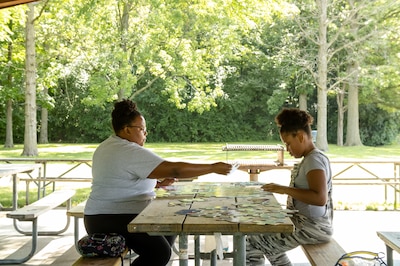 An adult on the left and a child on the right, both wearing light colored t-shirts sit at a picnic table with green trees in the background.