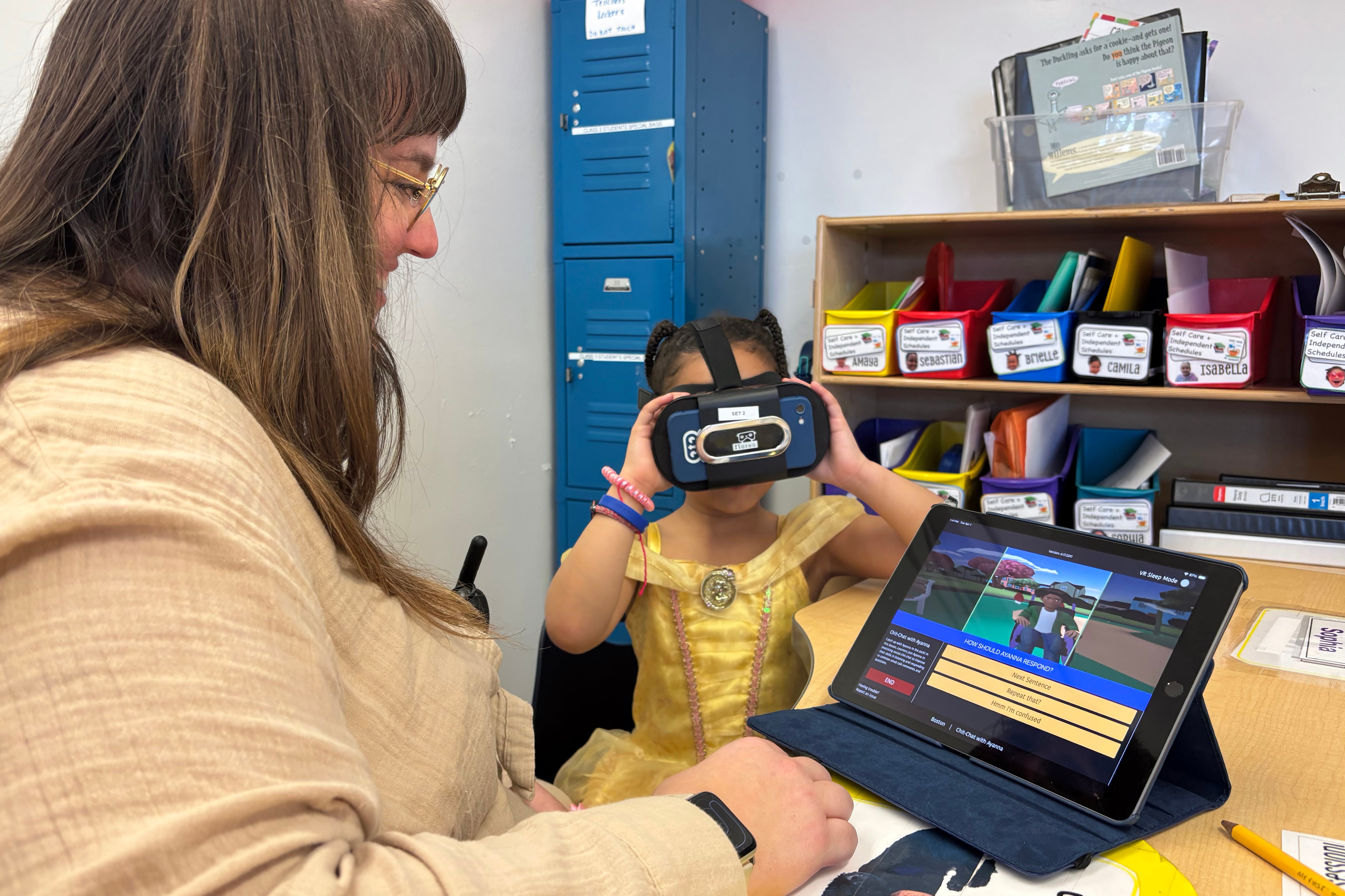 A teacher sits at the left of a young student wearing virtual realty goggles in a classroom.