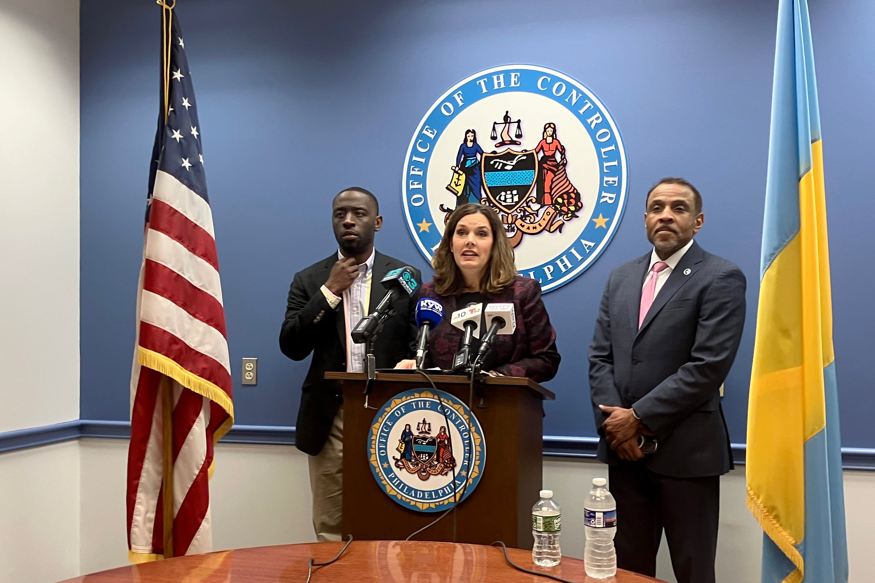 A woman stands between two men and two flags, behind a podium and in front of the city seal in a conference room.