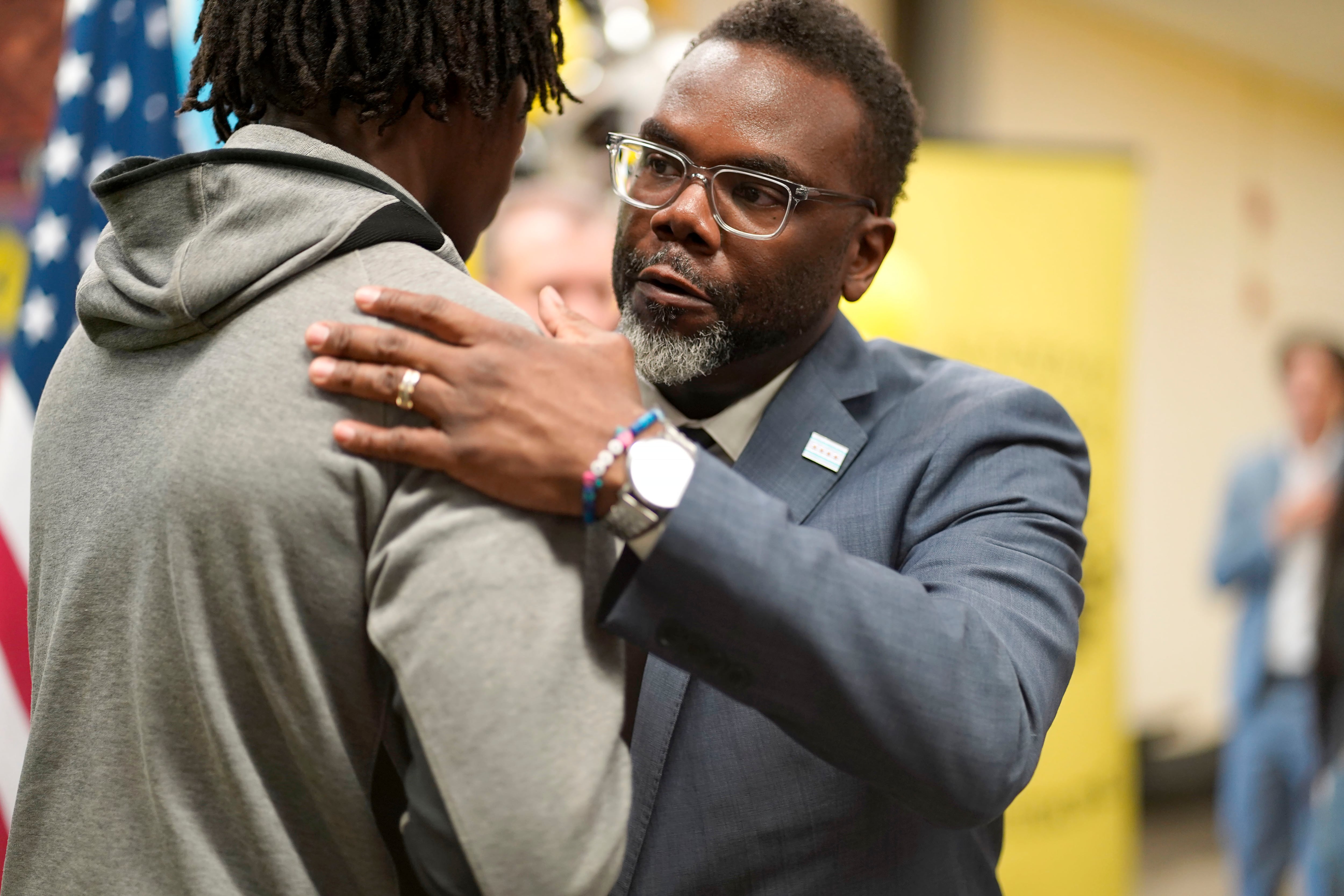 Chicago Mayor Brandon Johnson, wearing glasses and a gray suit jacket, hugs a teen in a gray sweat jacket. There’s an American flag and one perso in the background.