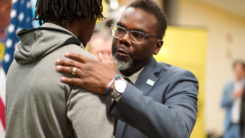 Chicago Mayor Brandon Johnson, wearing glasses and a gray suit jacket, hugs a teen in a gray sweat jacket. There’s an American flag and one perso in the background.
