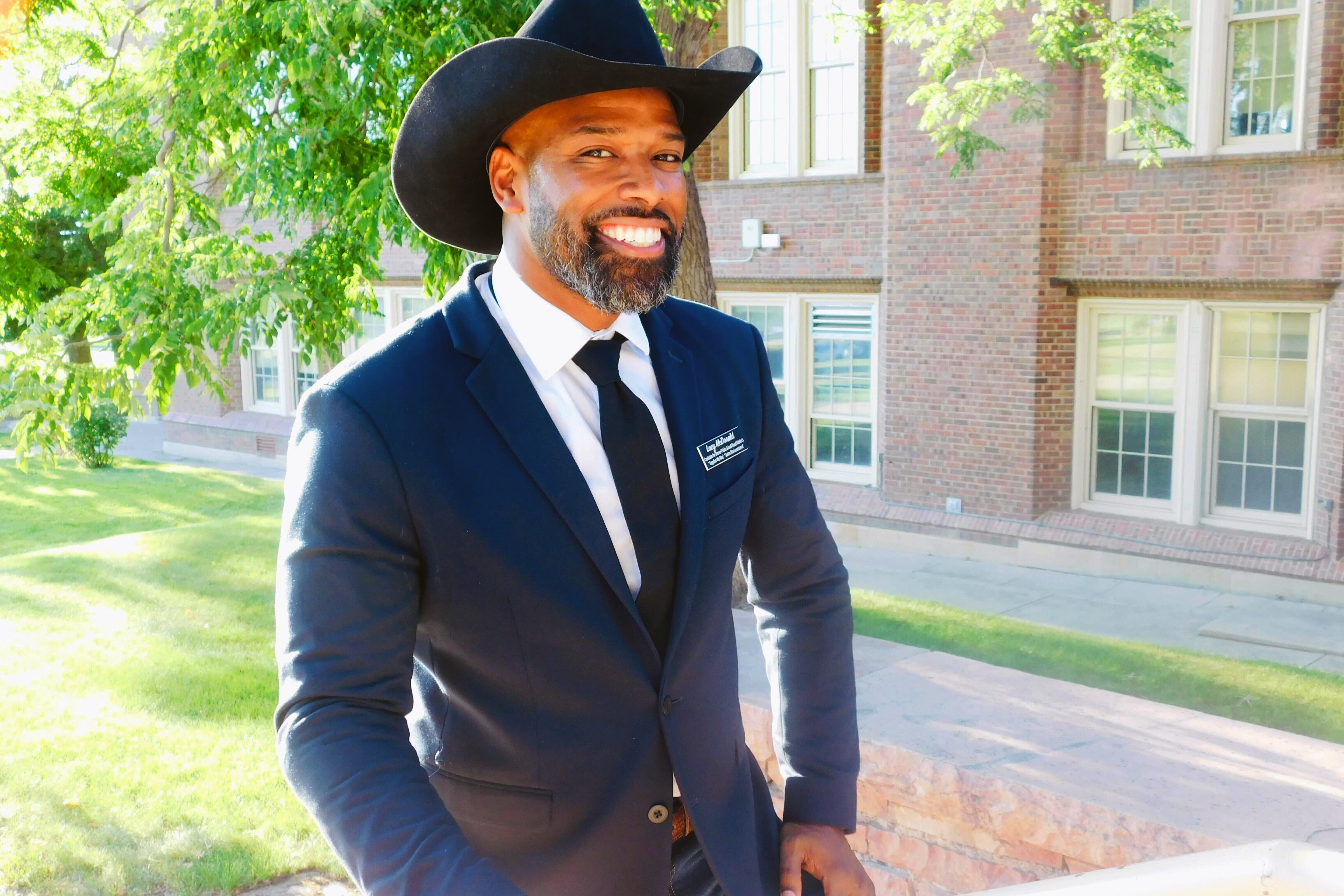 Images shows Lacy McDonald posing in front of a school. He is a Black man wearing a dark suit and a cowboy hat. He has a neatly trimmed beard and a huge smile on his face.