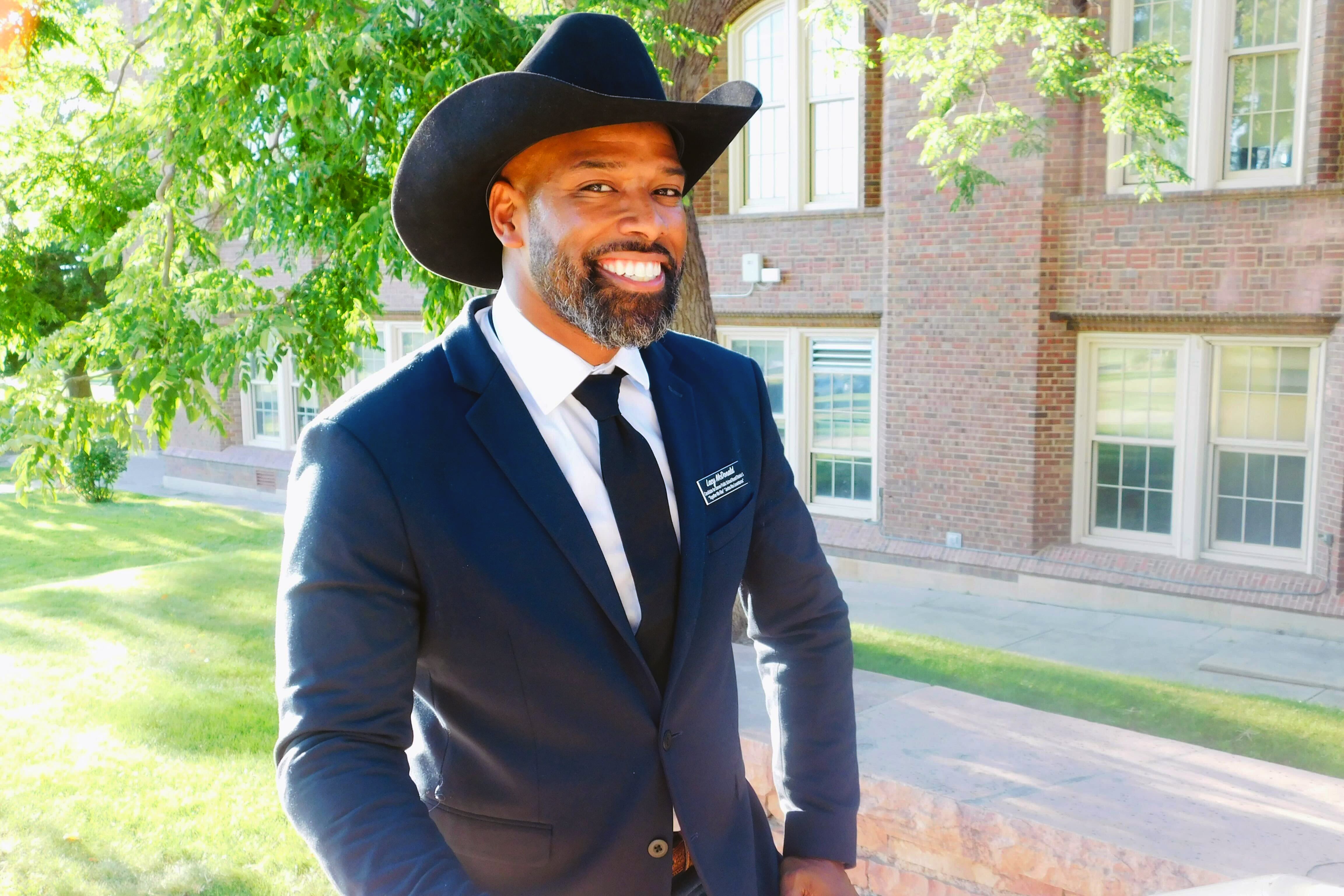 Images shows Lacy McDonald posing in front of a school. He is a Black man wearing a dark suit and a cowboy hat. He has a neatly trimmed beard and a huge smile on his face.