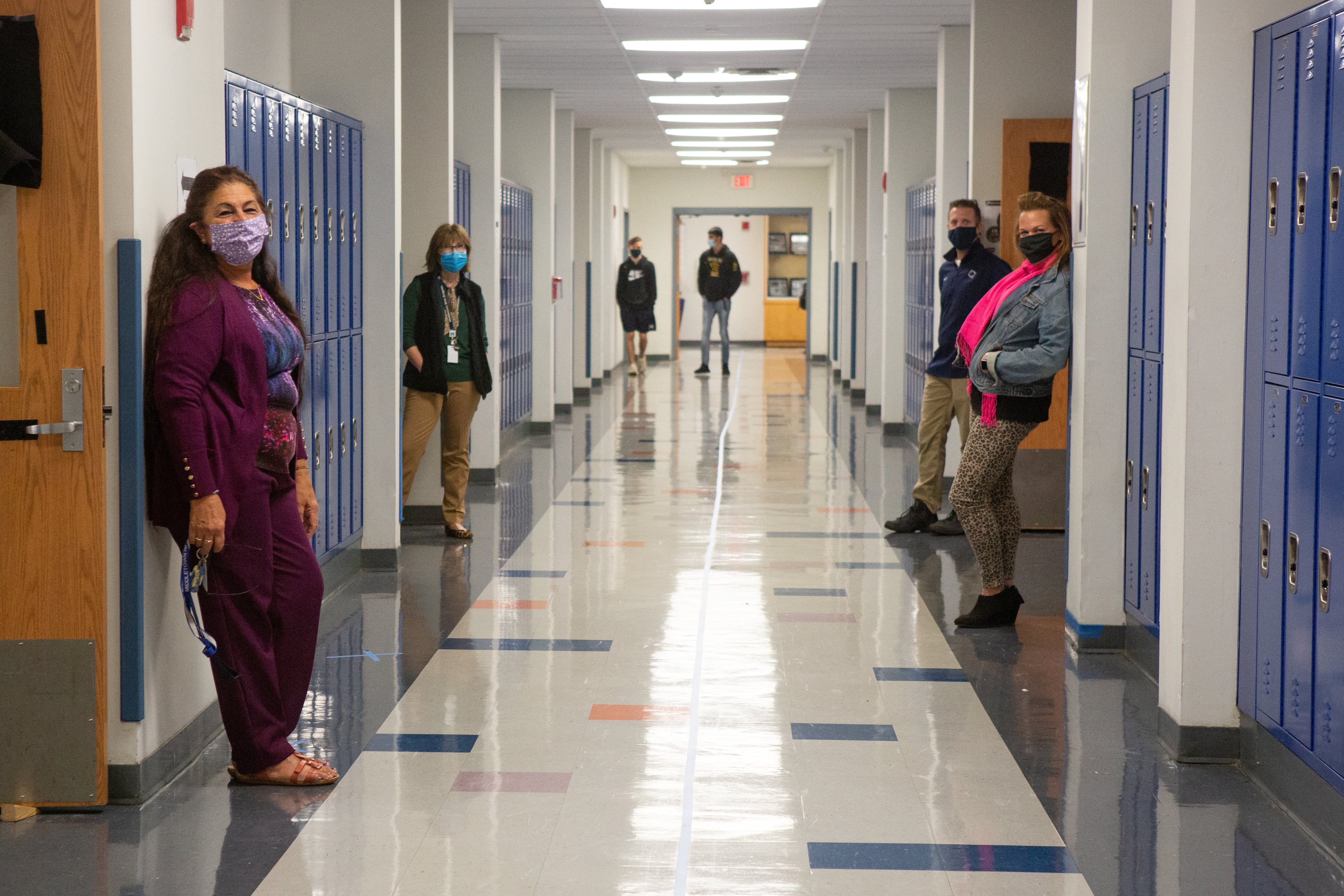 Masked teachers stand next to room doors in a long school hallway.