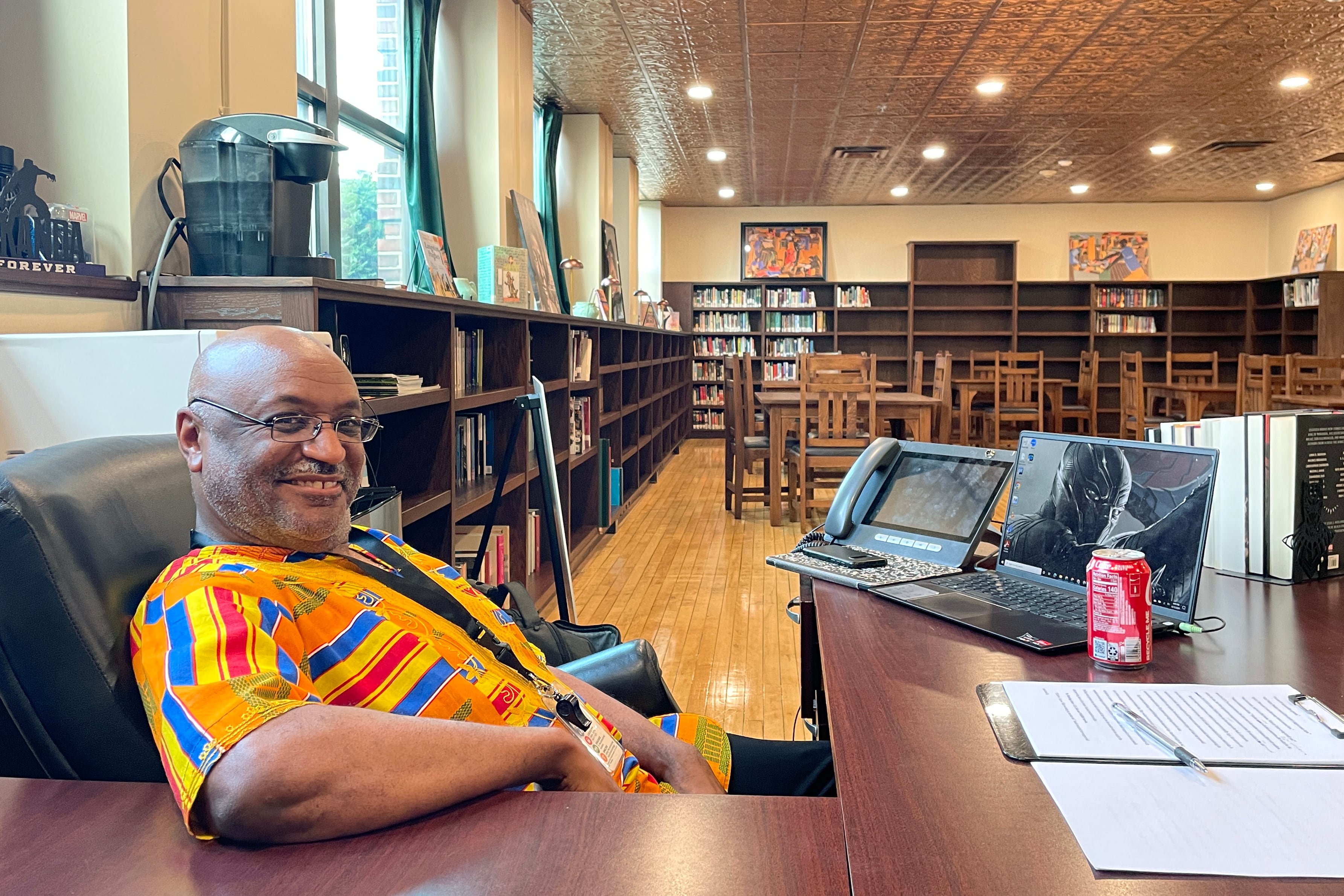 A man wearing glasses and a colorful shirt sits behind a wooden desk and smiles for a portrait in a room at the library with bookshelves and books lining the wall in the background.