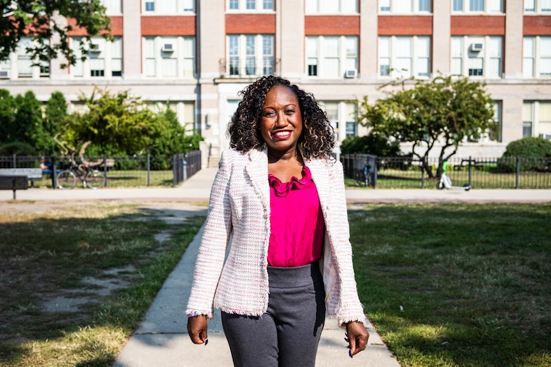 A woman with a white jacket and pink top stands in front of a building.