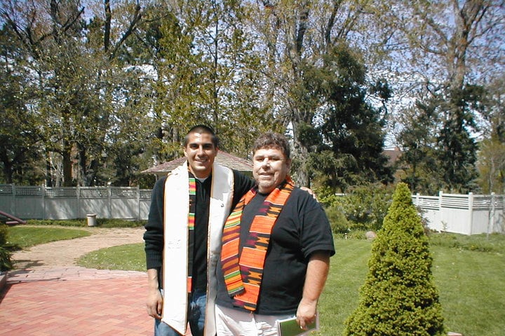 Two men dressed in black shirts and multi-colored graduation sashes stand in front of a courtyard with trees, a patio and grass.