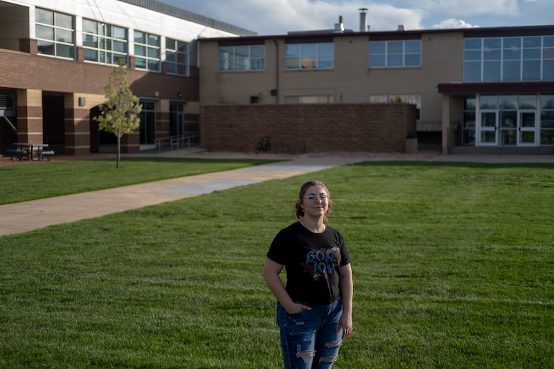 A young woman in a black t-shirt and jeans stands on grass in front of a building.