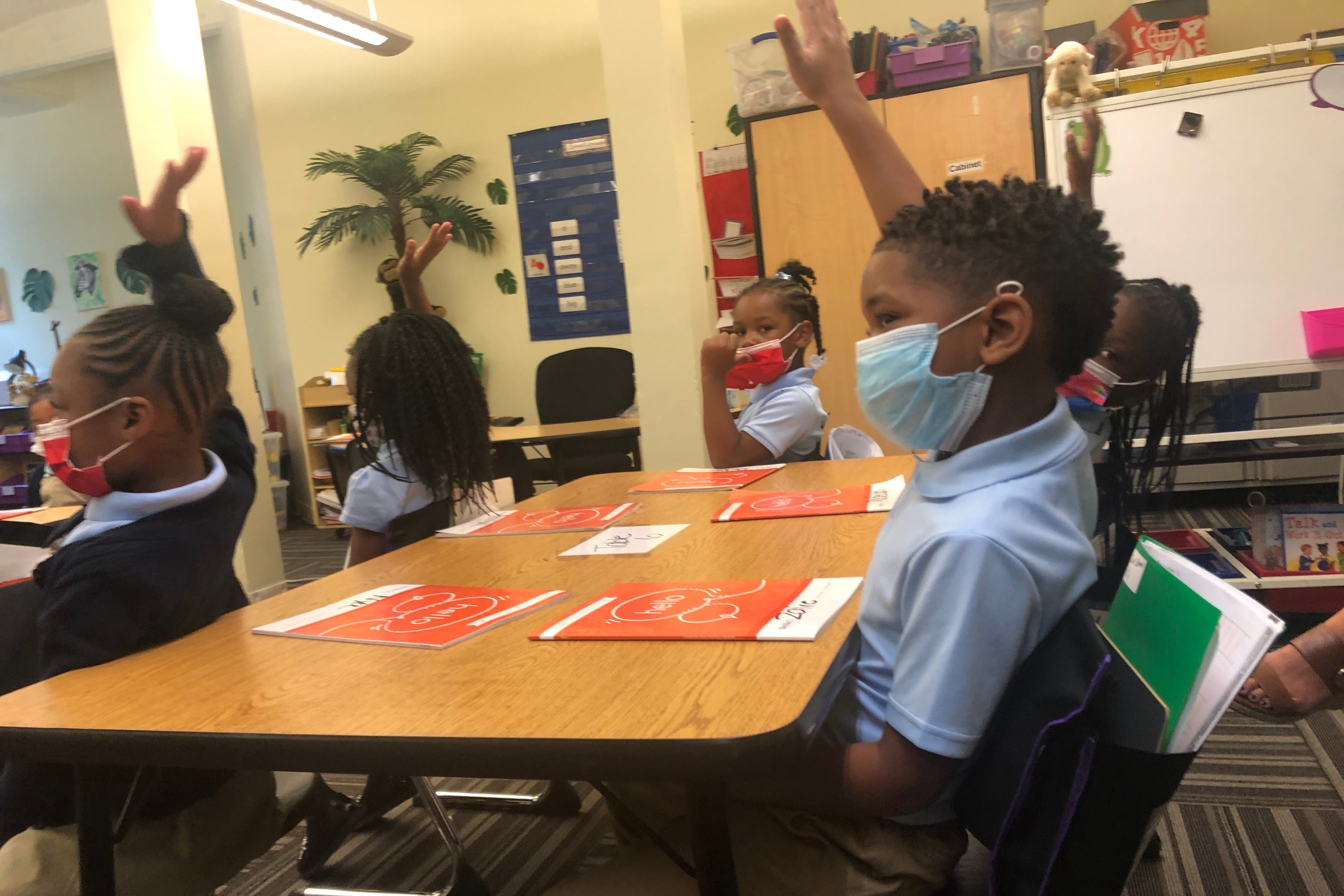 Kindergarten students wearing blue shirts and wearing face masks sit at tables and raise their hands during a lesson.