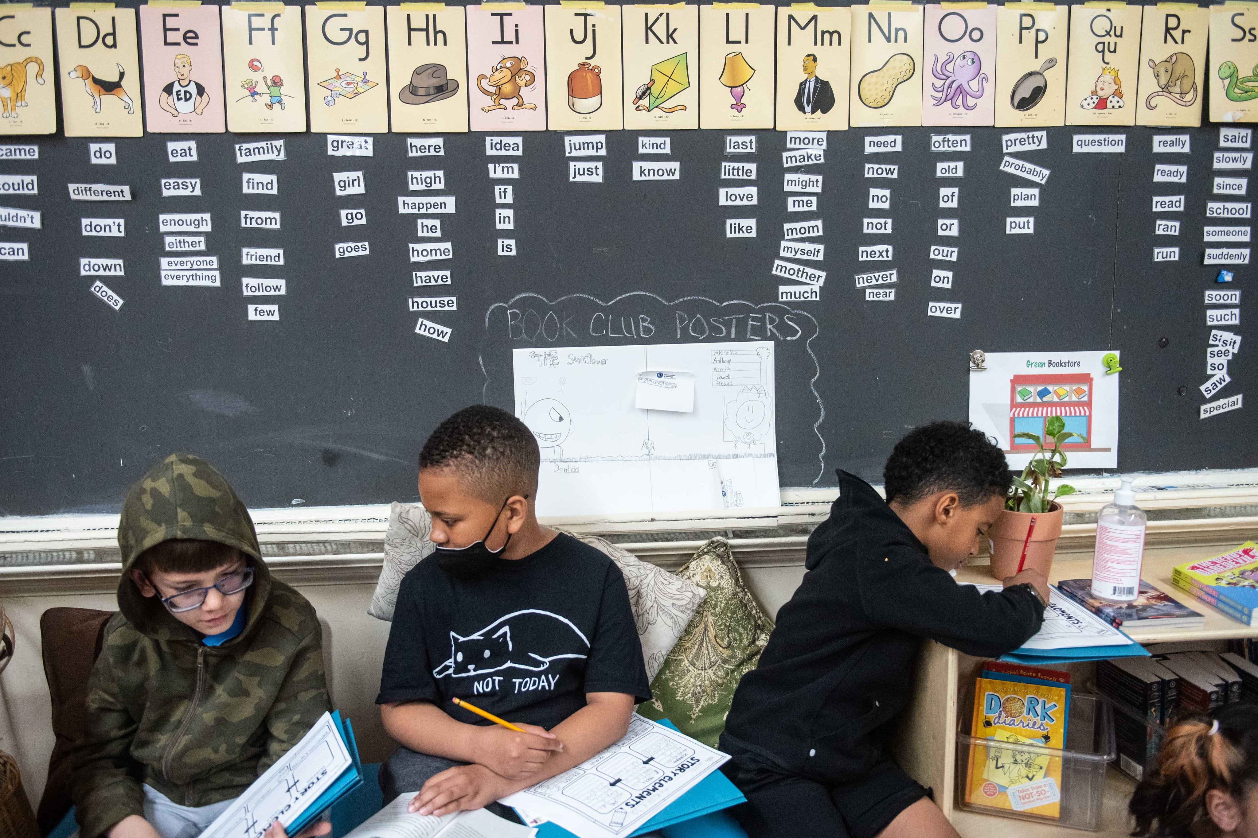 Three students sit with work in front of a chalkboard.