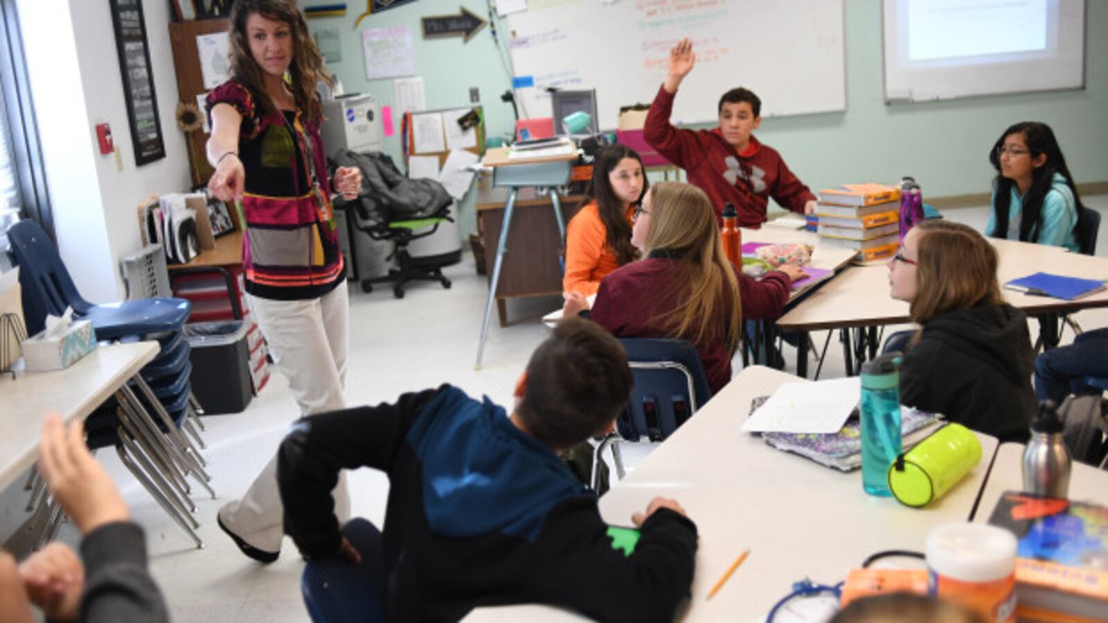 Stephanie Wujek teaches science at Wiggins Middle School, on April 5, 2017 in Wiggins, Colorado. A woman with shoulder-length brown hair stands at the front of the classroom and points to a student. Other students sit at tables, and one in a red hoodie raises their hand.
