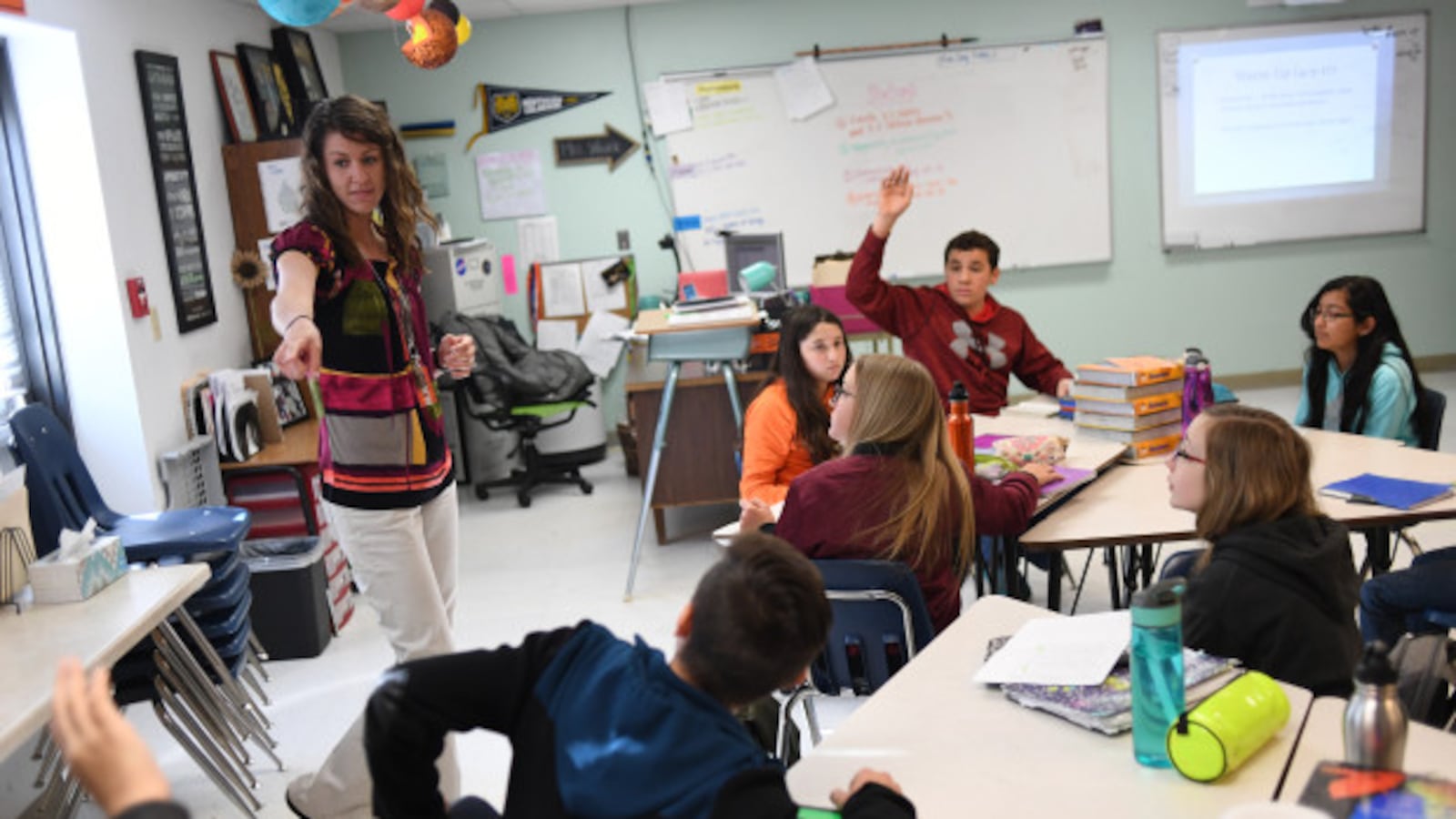 Stephanie Wujek teaches science at Wiggins Middle School, on April 5, 2017 in Wiggins, Colorado. A woman with shoulder-length brown hair stands at the front of the classroom and points to a student. Other students sit at tables, and one in a red hoodie raises their hand.