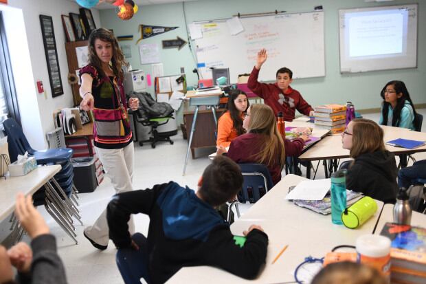 Stephanie Wujek teaches science at Wiggins Middle School, on April 5, 2017 in Wiggins, Colorado. A woman with shoulder-length brown hair stands at the front of the classroom and points to a student. Other students sit at tables, and one in a red hoodie raises their hand.
