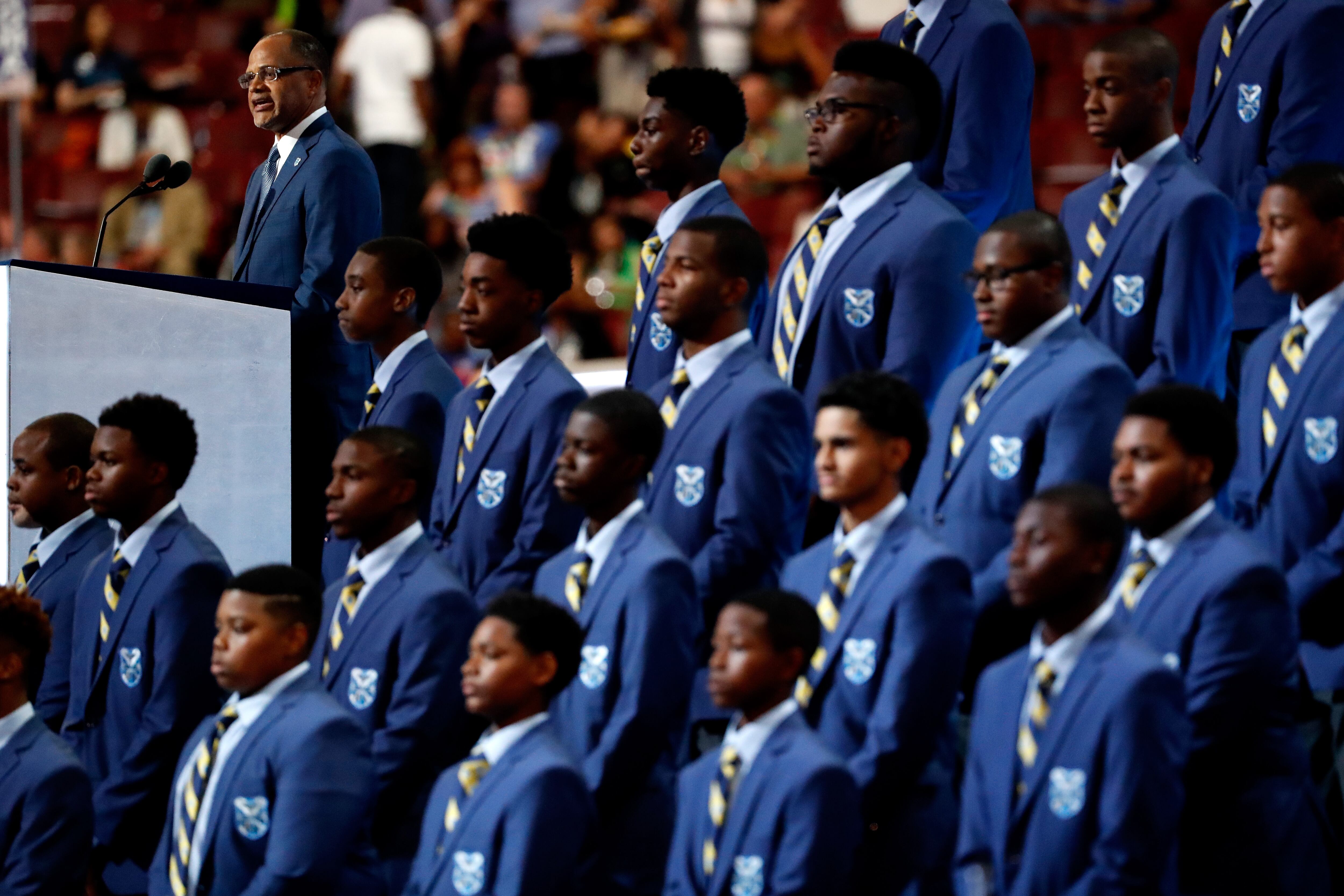 Rows of students, wearing blue Eagle Academy jackets and ties, stand next to a podium where incoming NYC Chancellor David Banks is speaking.