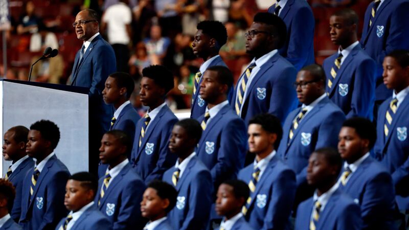 Rows of students, wearing blue Eagle Academy jackets and ties, stand next to a podium where incoming NYC Chancellor David Banks is speaking.