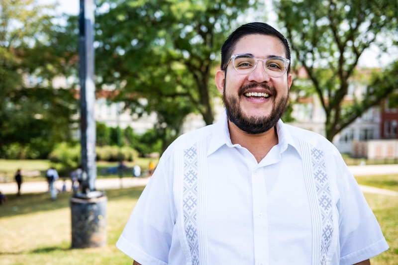A man wearing a white dress shirt stands in front of a green background outside.