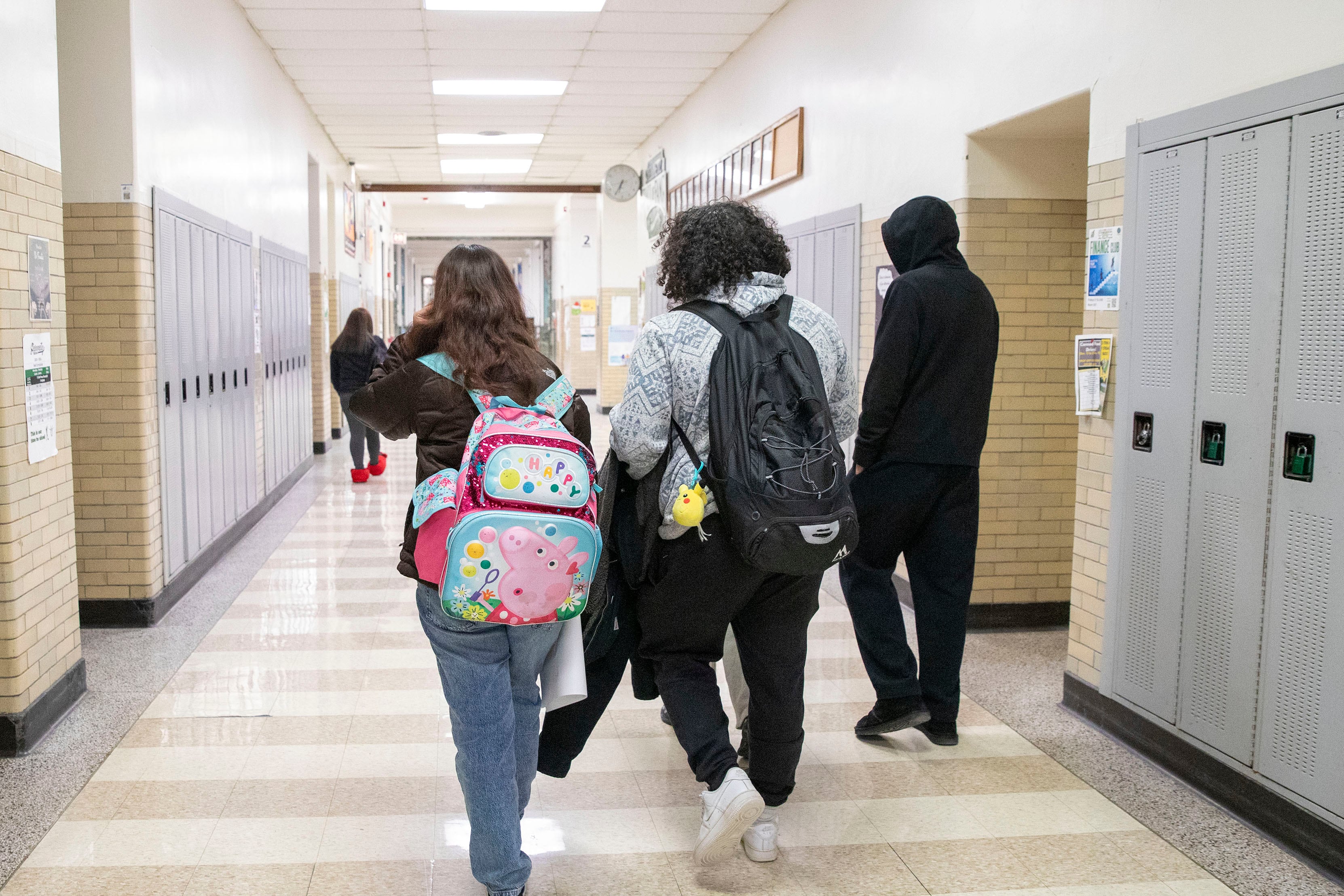 A photograph of high school students walking down a hallway with lockers on each side.
