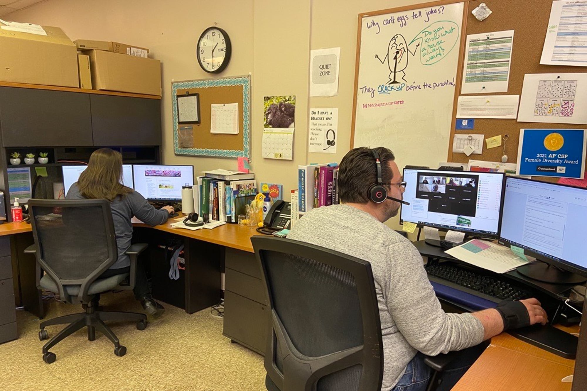Two teachers sit at computers teaching virtual classes with headphones on.