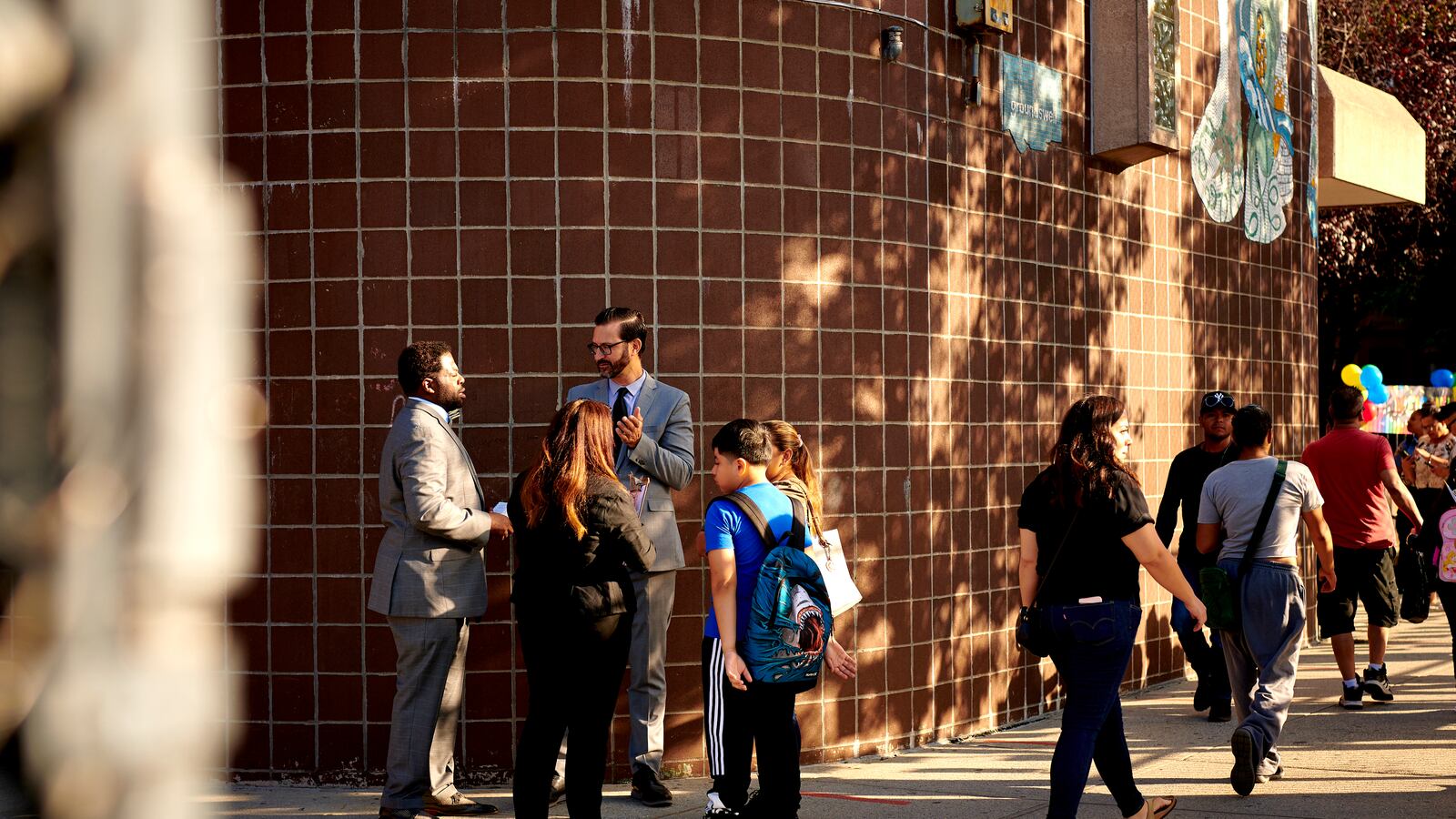 School administrators speak with a family on the first day of school as other parents walk by them outside a brick school building.