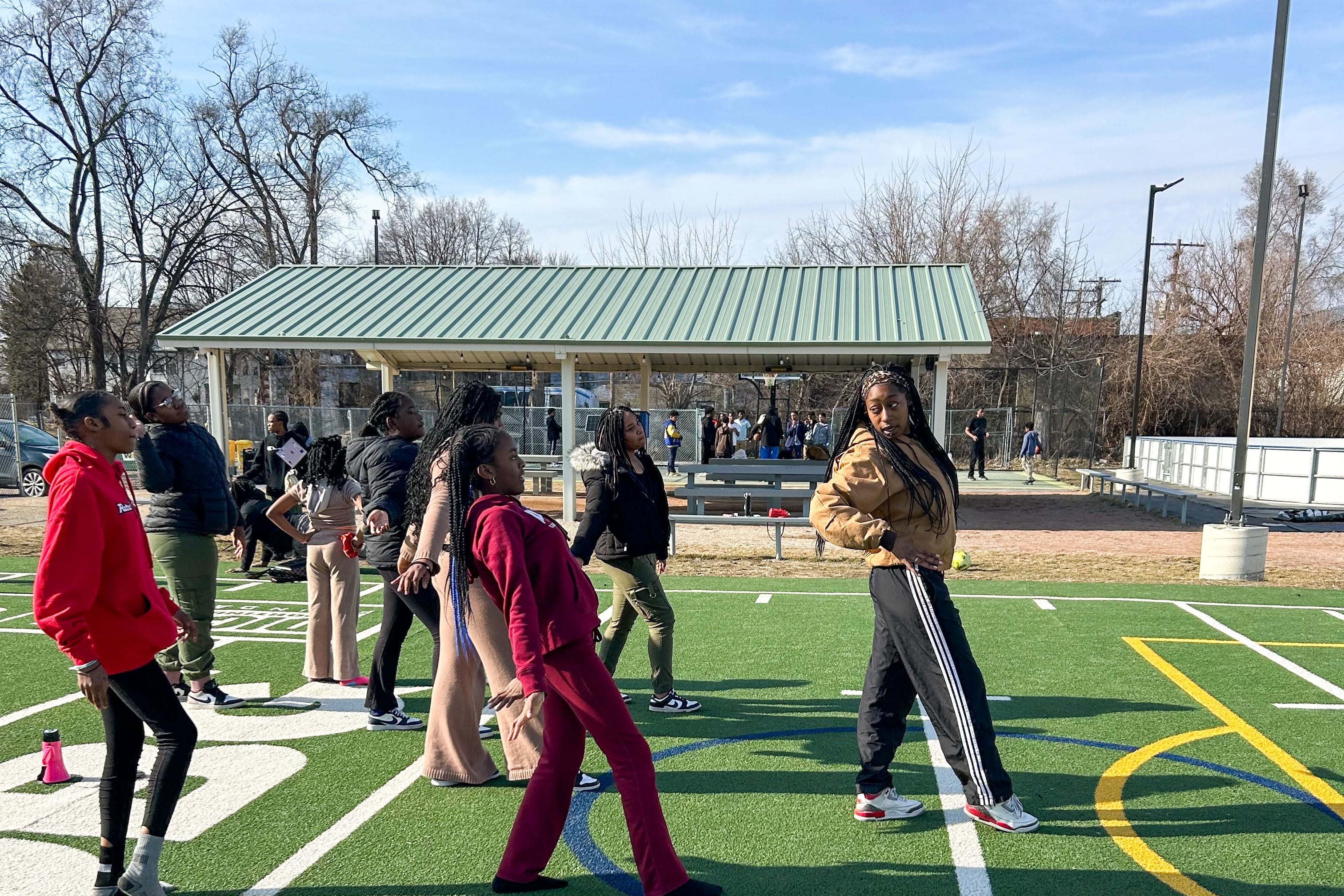 A group of high school students practice a dance routine outside on a football field.