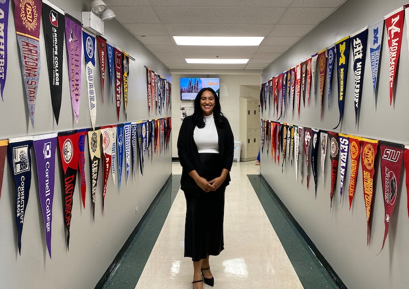 A woman in a black skirt and white shirt stands for a photo in a school hallway lined with college pendants.